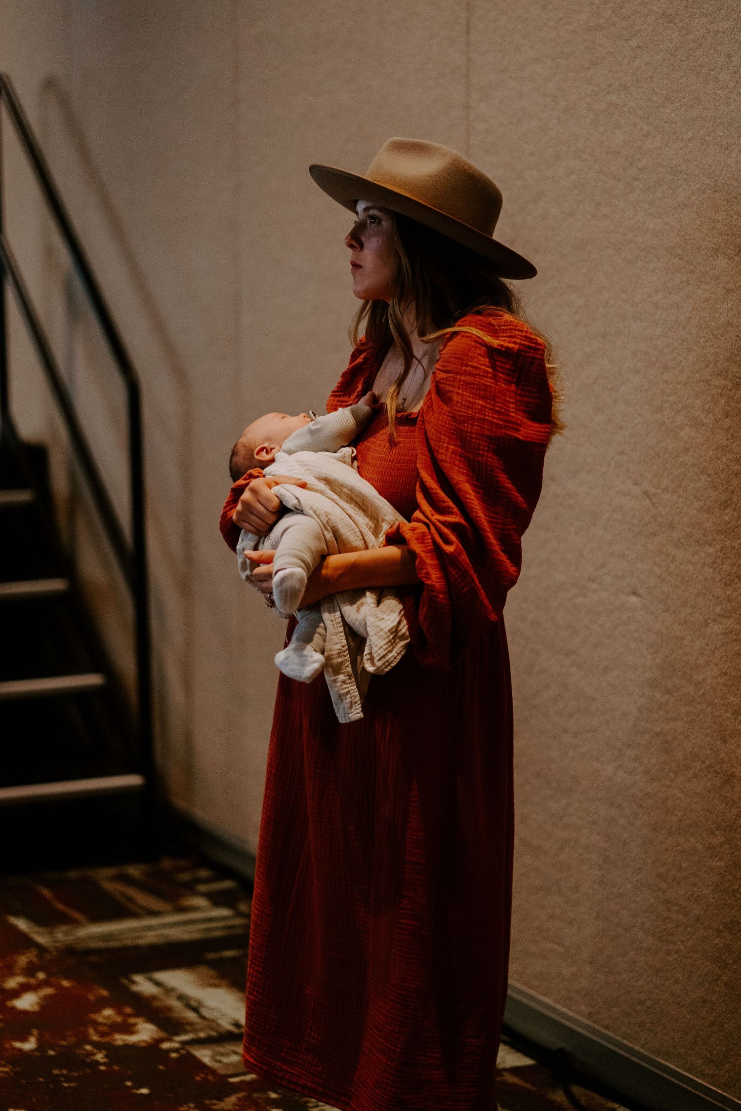 Woman in a wide-brimmed hat and rust-red dress cradles a sleeping newborn beside a staircase.