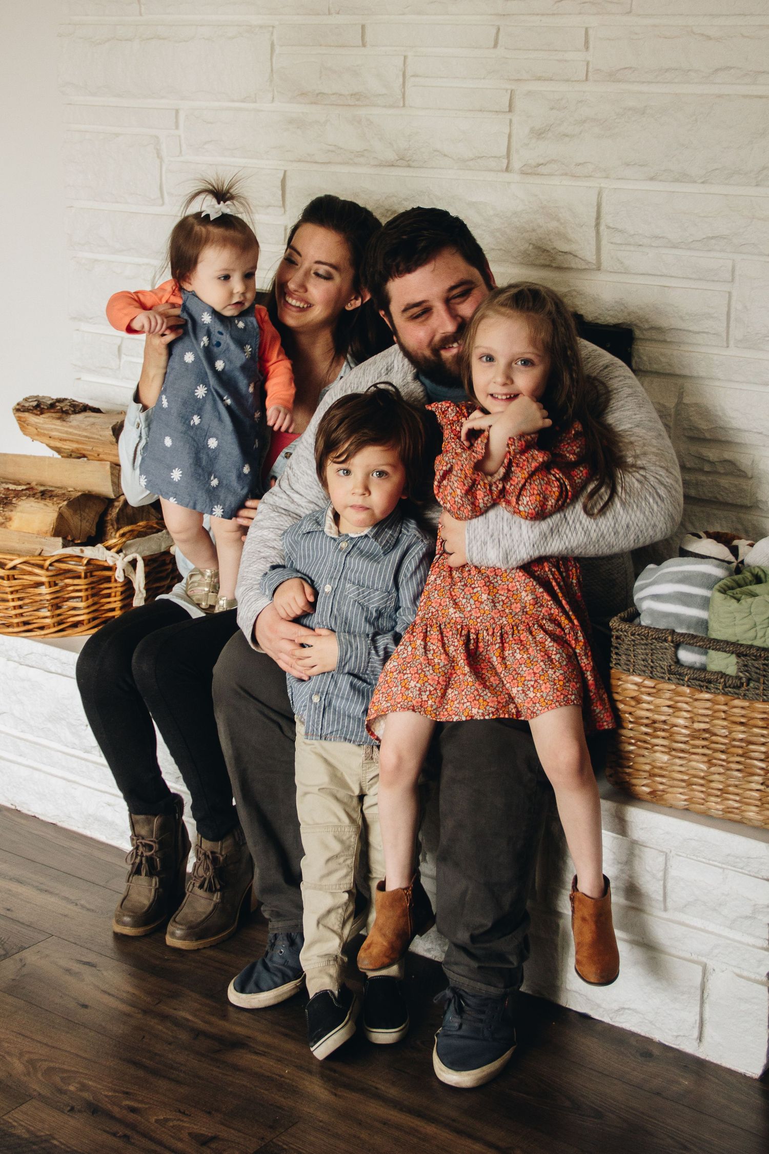 A happy family portrait of two parents and three young children cuddled together on a white brick hearth beside woven baskets.