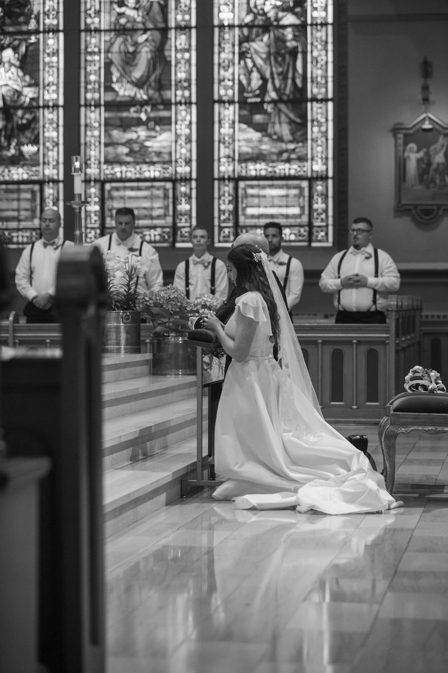 A black and white photo of a bride in a long veil kneeling in prayer at a catholic church as groomsmen stand beside stained glass windows.