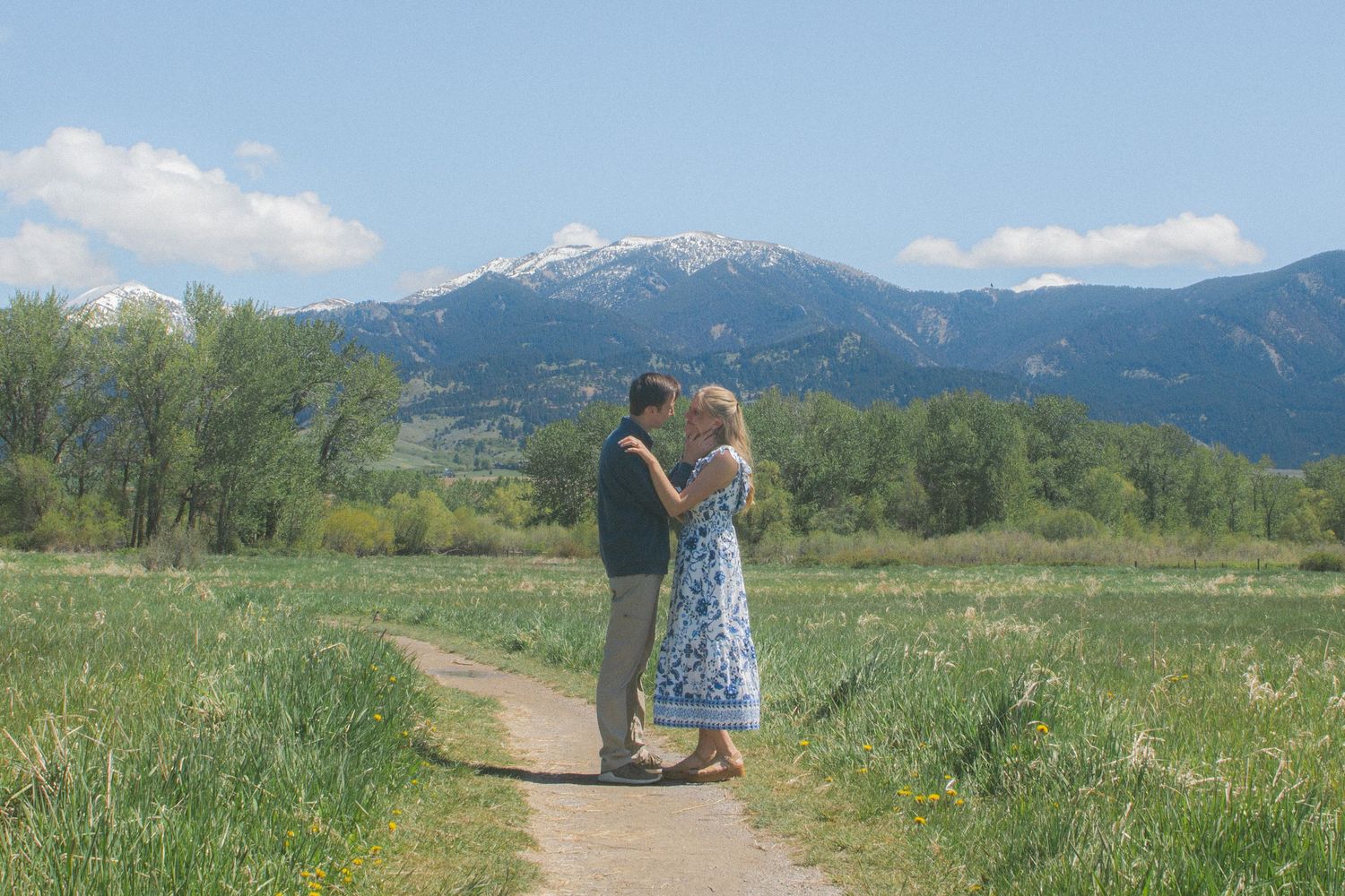 Couple embraces on a dirt path through a green meadow with snow-capped mountains and blue sky in the background.