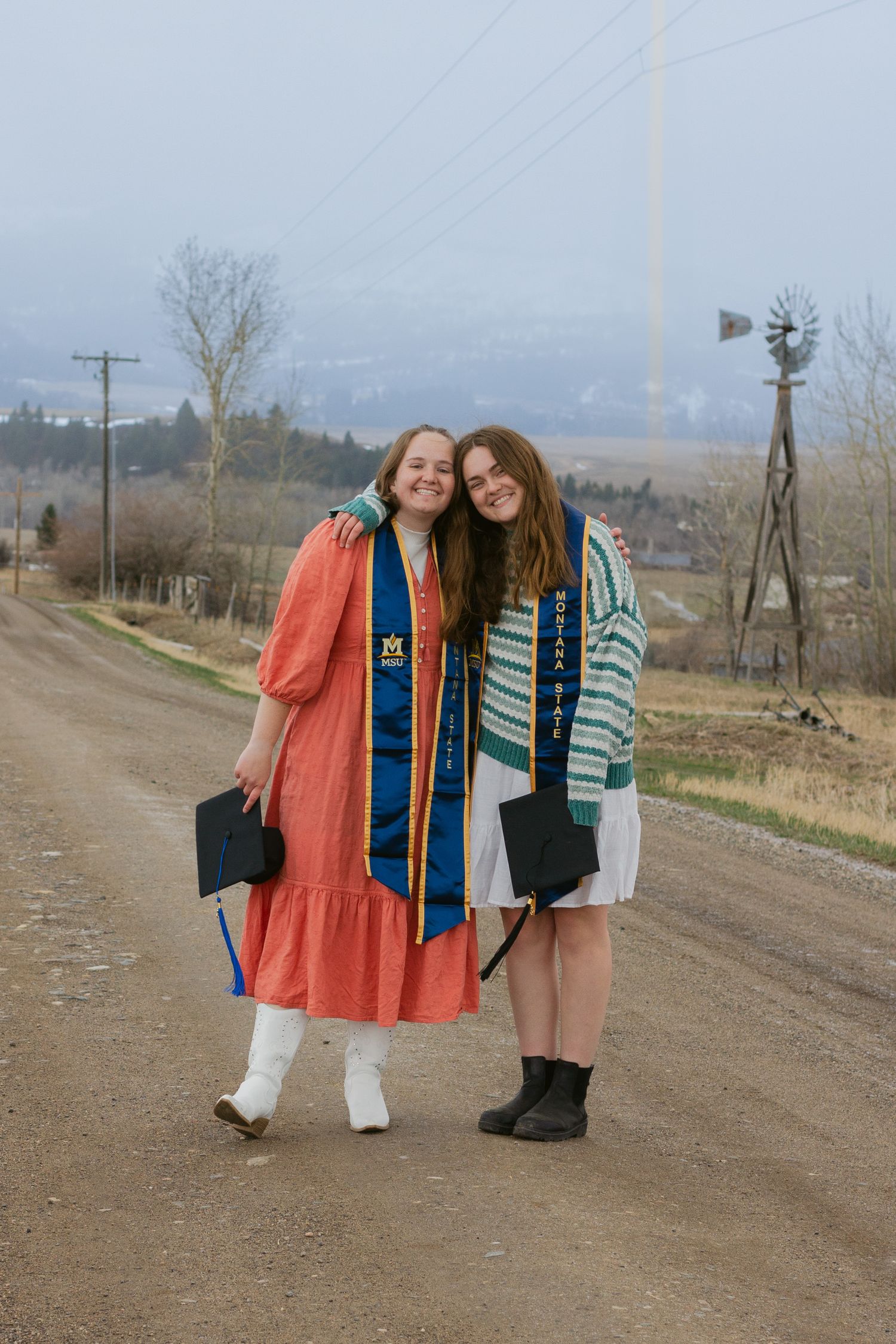 Two smiling graduates in colorful stoles hold caps and hug on a dirt country road with a windmill and misty hills.