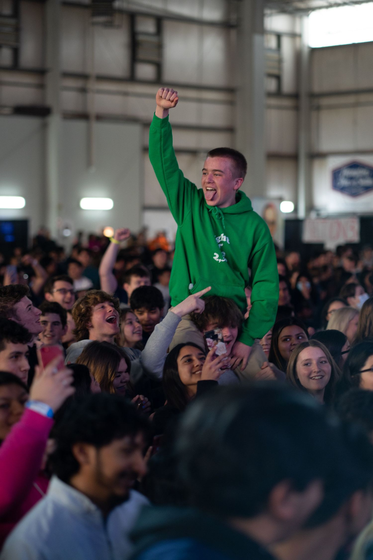 A teenager in a green hoodie sits on someone's shoulders with a raised fist, cheering in an energetic crowd at an indoor event.