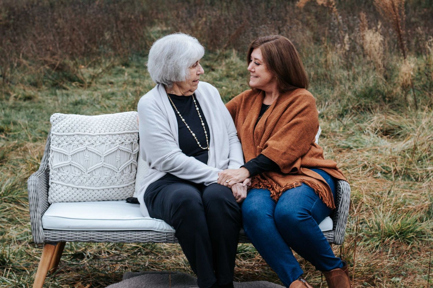 An older woman and a younger woman hold hands and smile while seated on a wicker bench in a grassy autumn field.