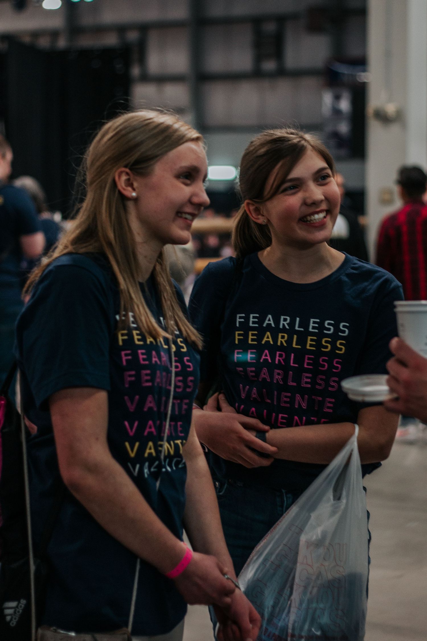 Two smiling girls in navy Fearless Valiente t-shirts chat happily while holding a clear bag at a youth event.