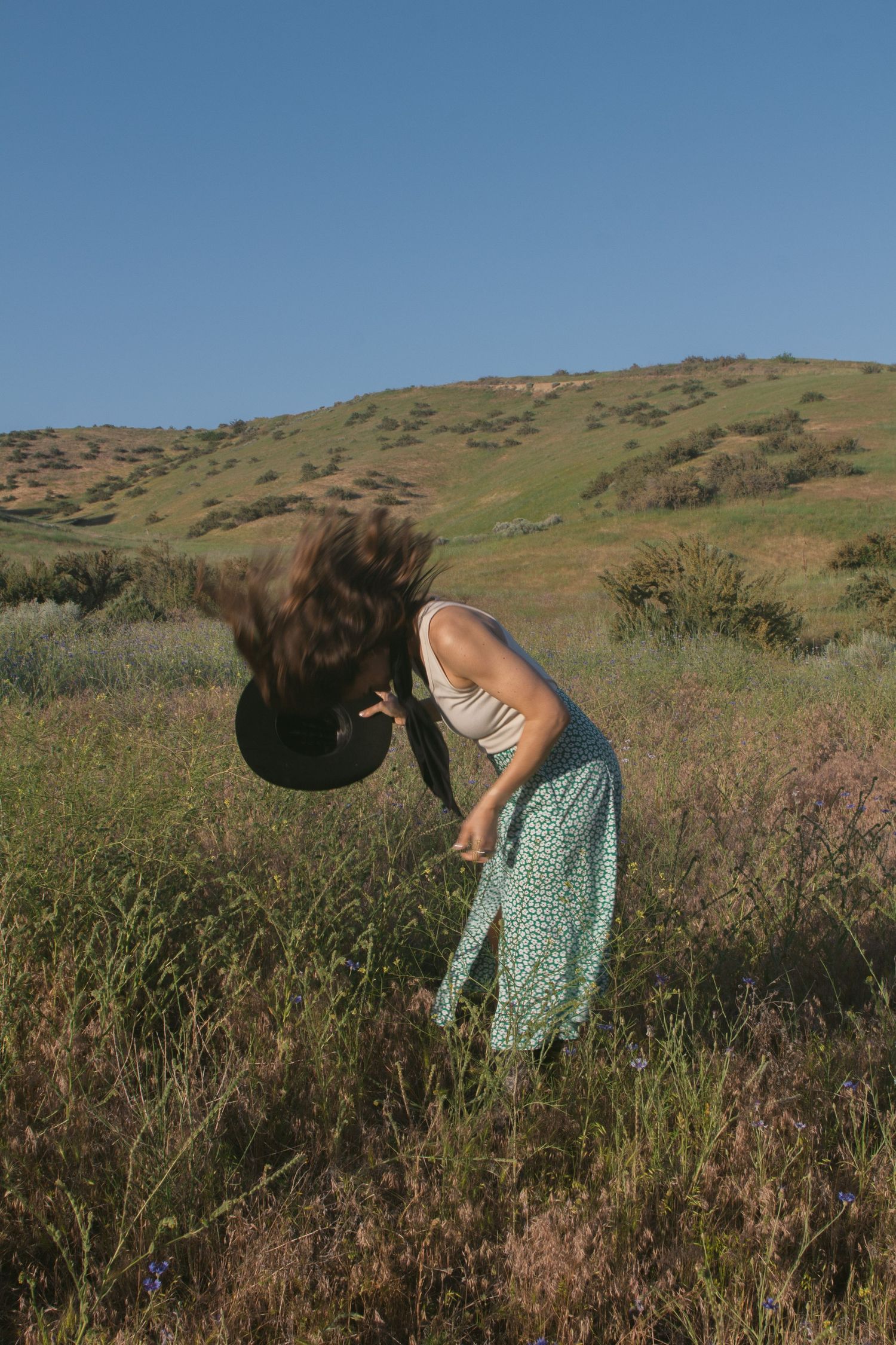 Woman in a green floral skirt flips her long brown hair forward while holding a black hat in a grassy hillside field.