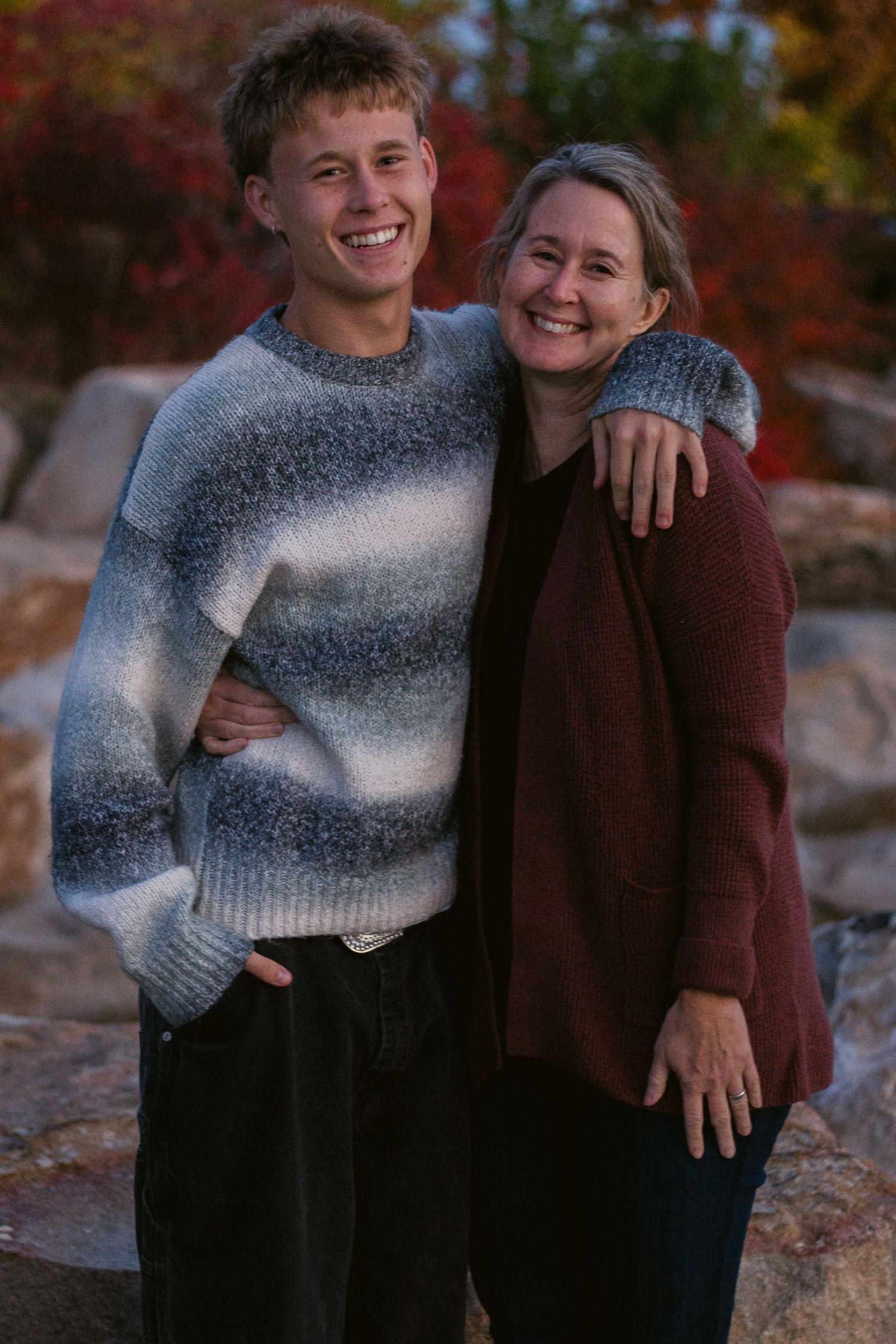 Smiling young man in a gray sweater hugs a woman in a maroon cardigan in front of rocks and autumn color.
