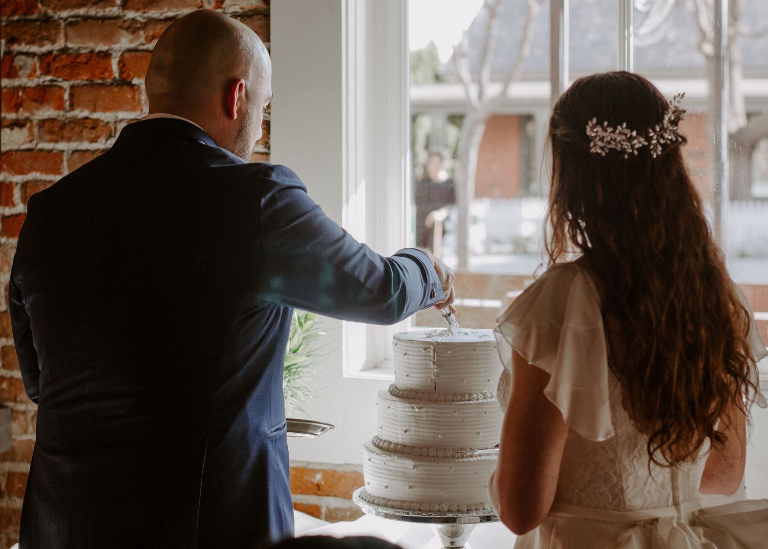 A bride with a floral hair piece and a groom in a navy suit cut a white tiered wedding cake beside a window and exposed brick wall.