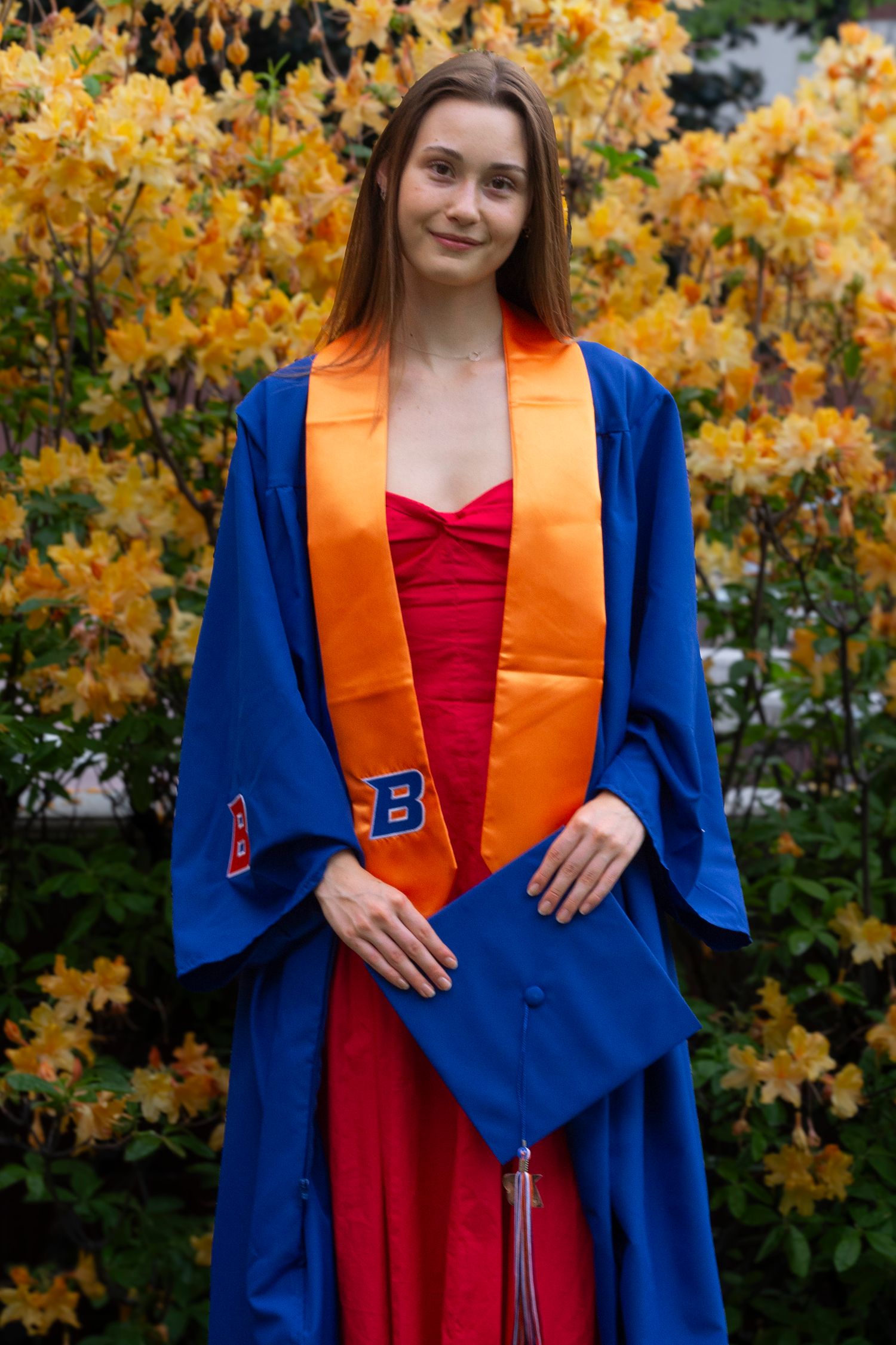 A smiling graduate in a blue gown and orange stole holds her cap while standing in front of bright yellow azalea blooms.