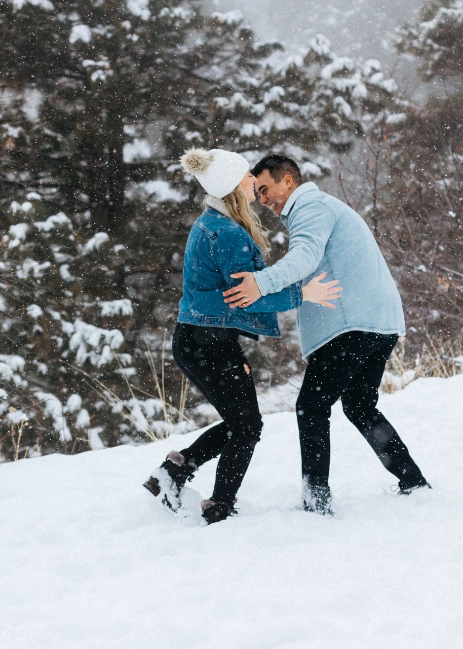 A couple in a denim jacket and light blue sweater embrace and laugh in deep snow beside snow-dusted pine trees.