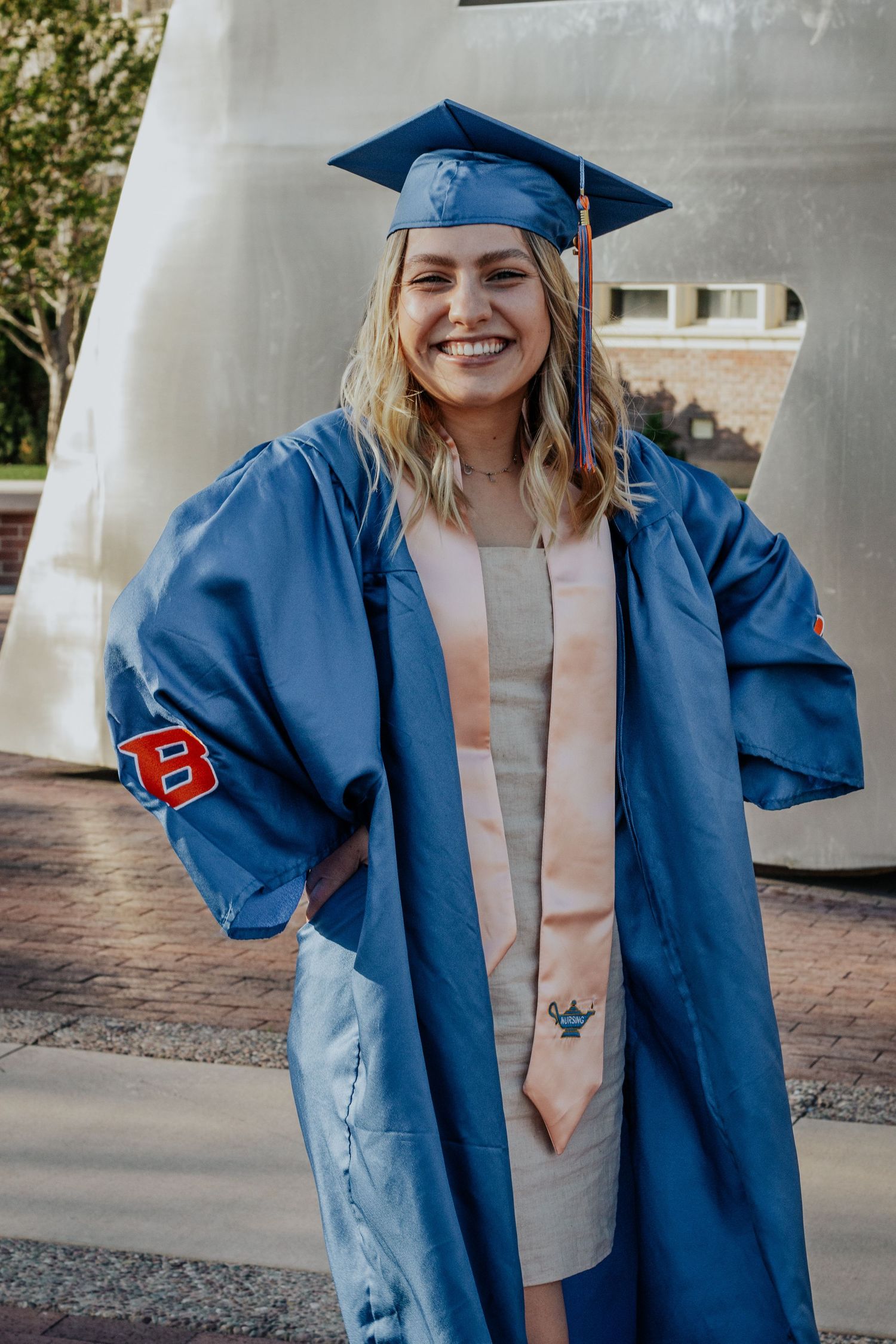 Cheerful graduate in a blue gown with a Boise State B on the sleeve and a peach stole poses in front of a large white sculpture.