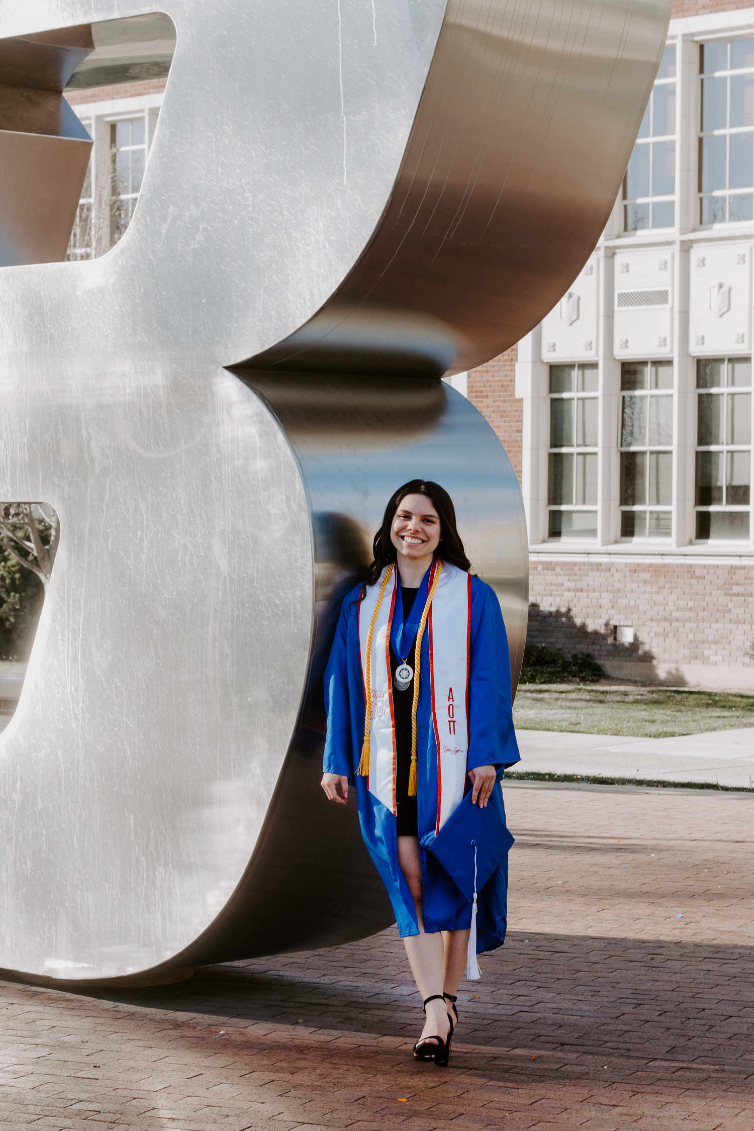 A smiling graduate in a blue gown and stole walks past a large silver sculpture on a brick campus.