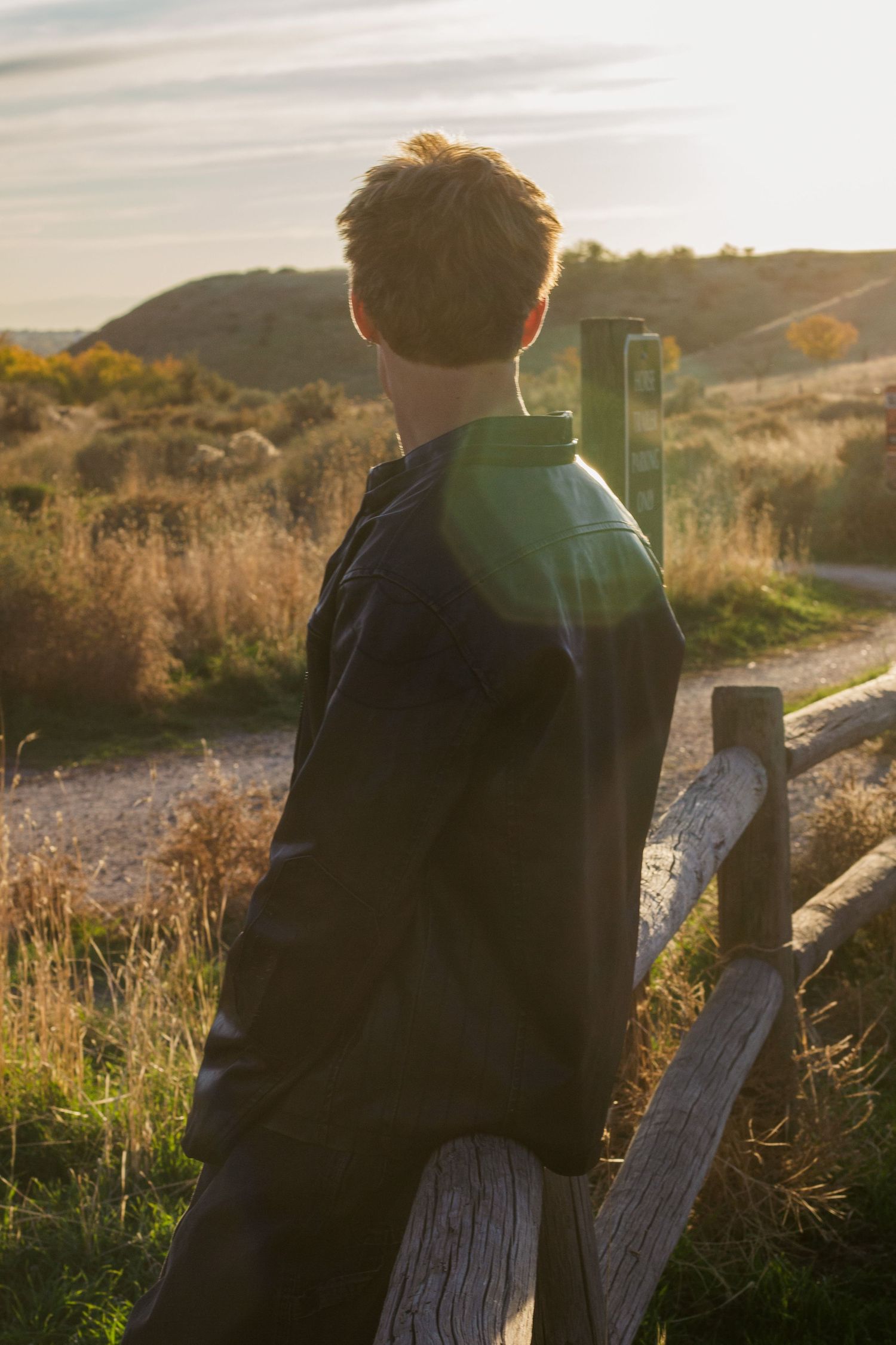 Young man in a dark jacket stands facing golden hills beside a rustic wooden fence at sunset.