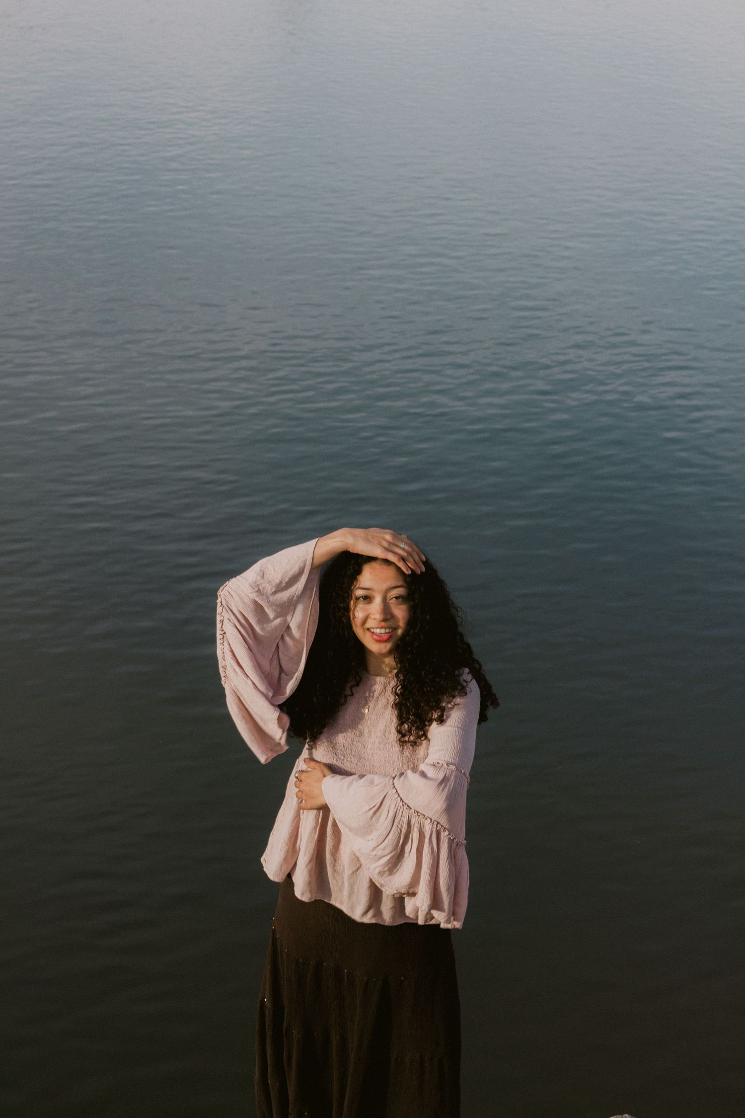 A smiling woman in a pink bell-sleeve blouse and brown skirt stands by dark rippling water with one hand resting on her head.