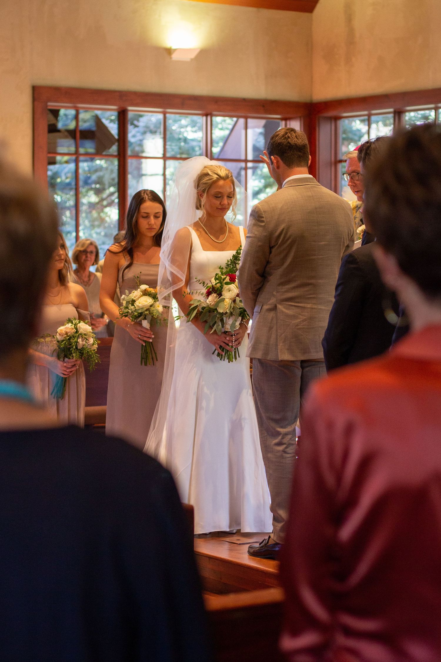 A bride in a white gown and veil holds a bouquet beside the groom and bridesmaids during a catholic wedding ceremony as guests look on.