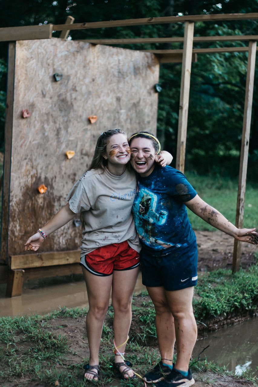 Two smiling, mud-splattered women hug in front of a wooden climbing wall at an outdoor camp obstacle course.