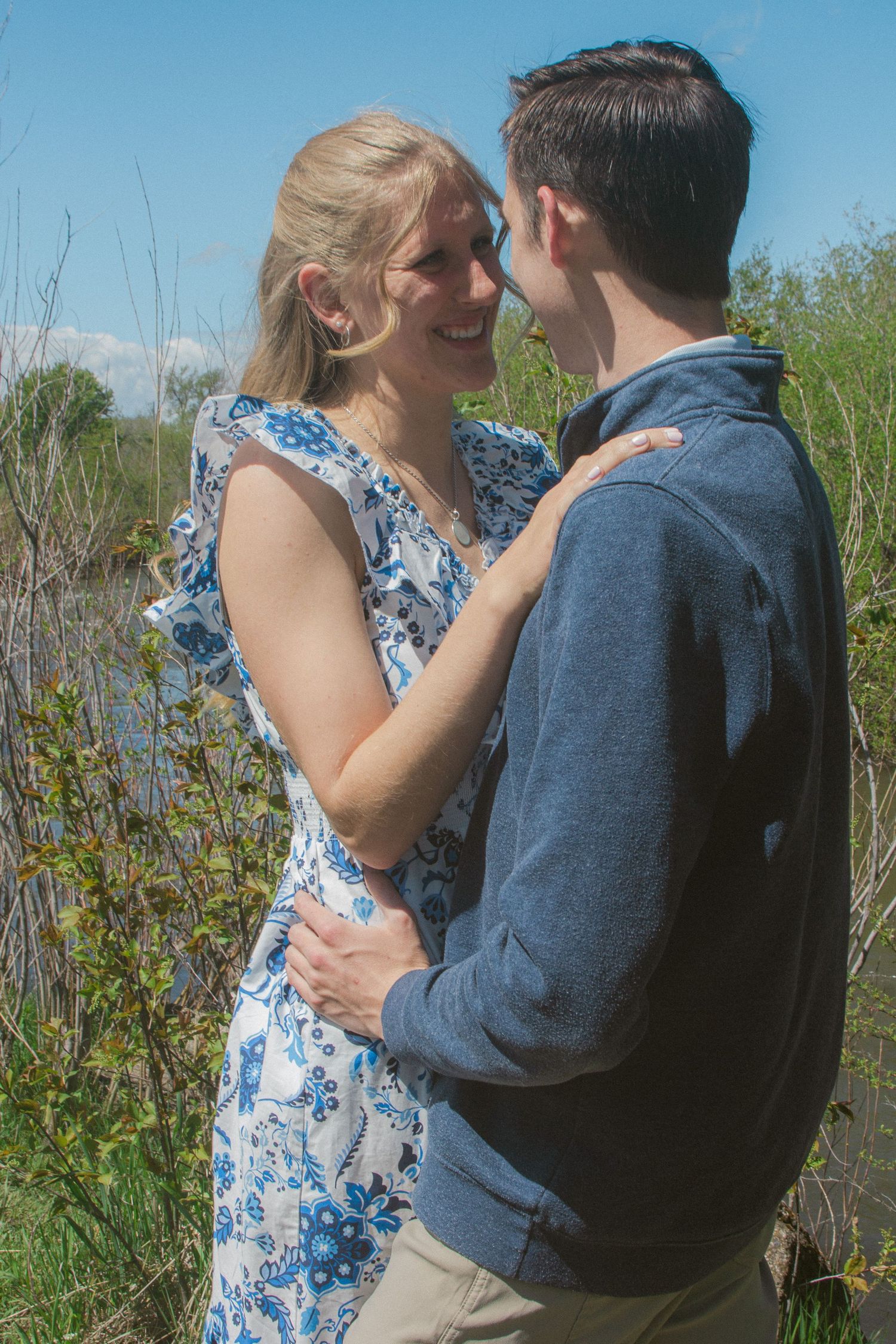 Couple in a blue floral dress and navy pullover embrace and smile at each other among green spring grasses.