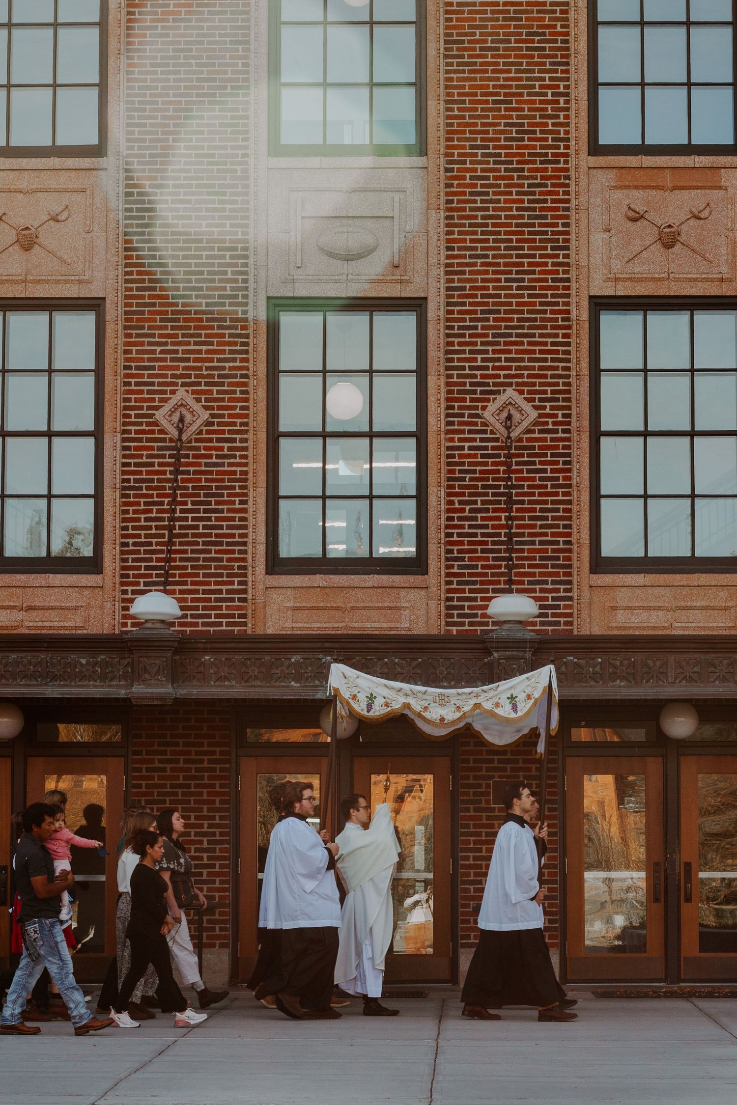 Clergy in white robes walk under a canopy in a Eucharistic procession past a brick church as parishioners follow on the sidewalk.