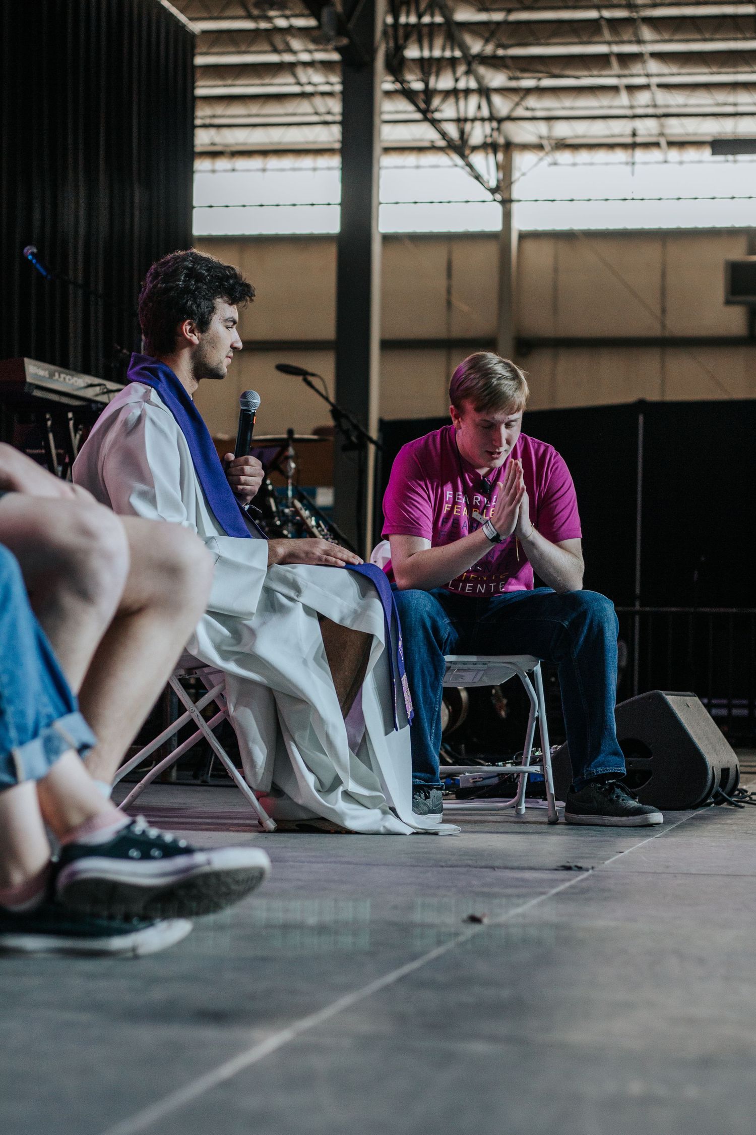 A young man in a white robe and blue stole speaks into a microphone beside a teen in a pink shirt with folded hands.