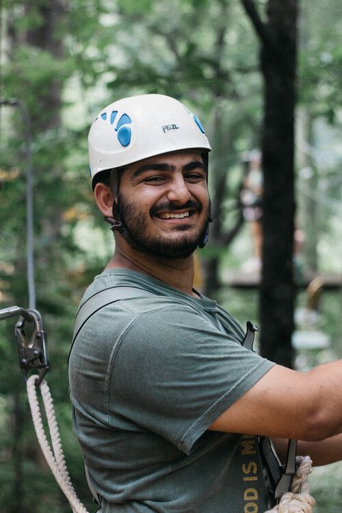 A smiling bearded man in a white helmet and harness prepares for a zipline or high ropes course amid tall forest trees.