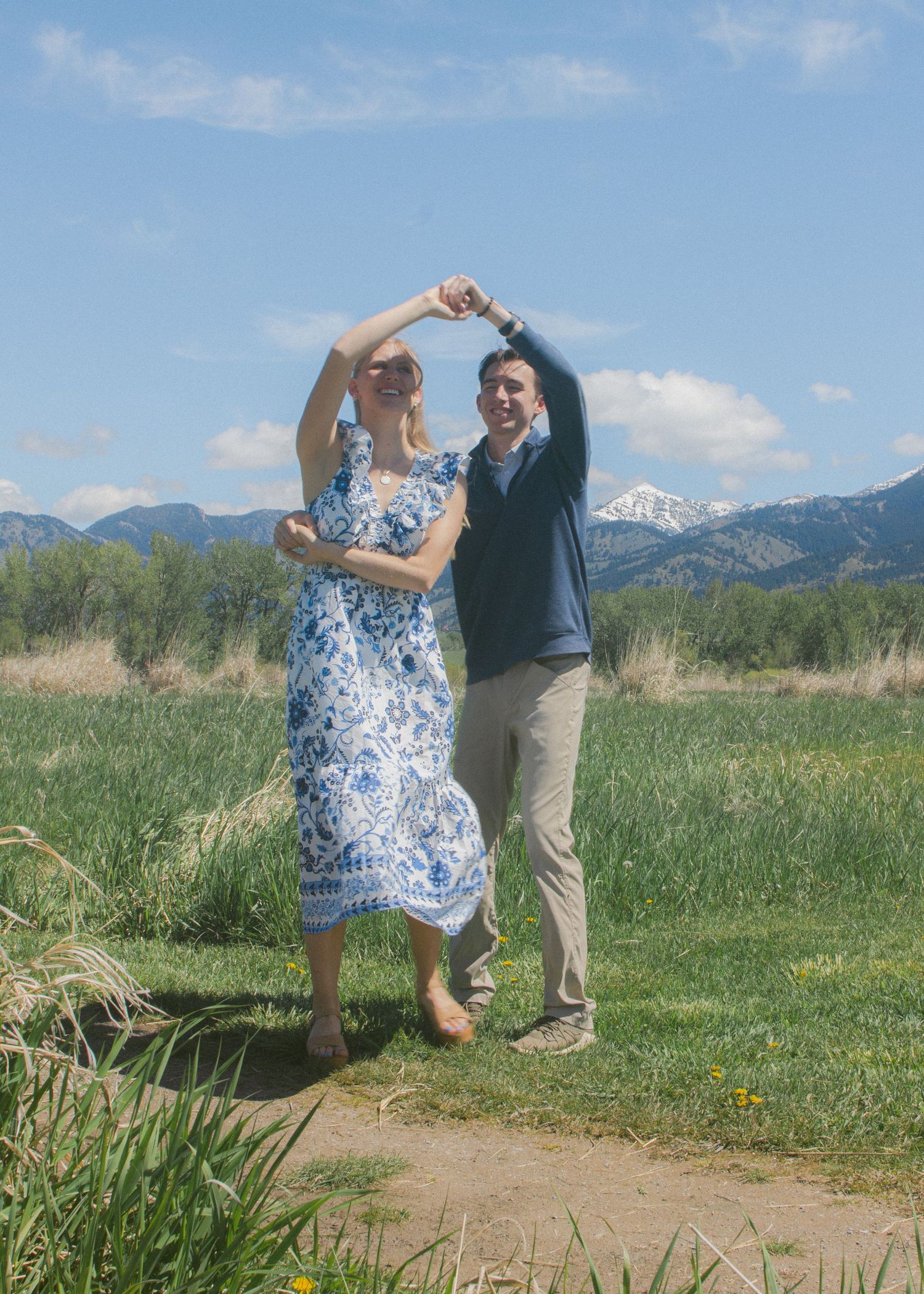 Couple dancing in a grassy field with snow-capped mountains behind, she twirls in a blue floral dress under a bright sky.