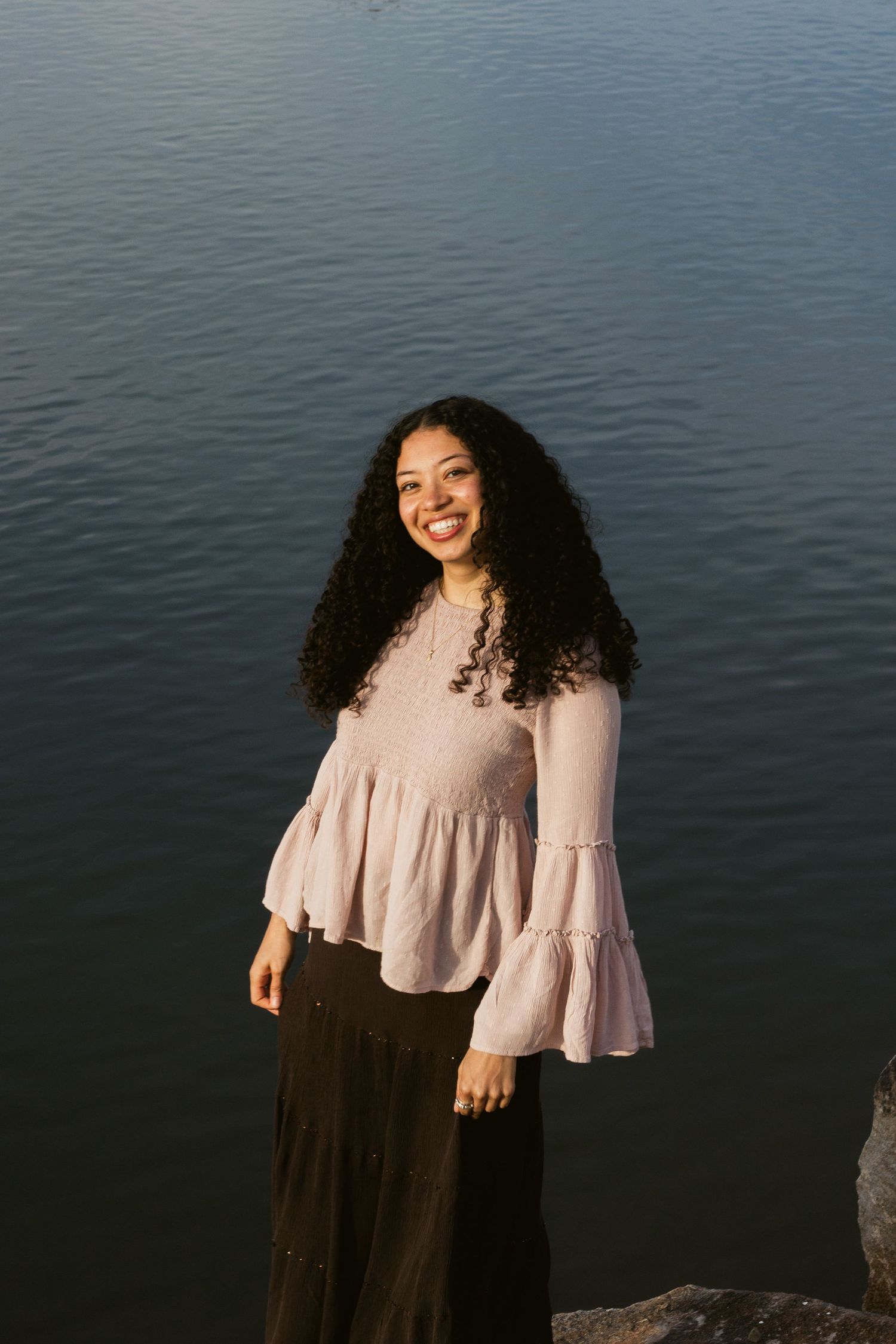 A smiling woman with long curly hair stands on rocks by the water in a pink ruffled blouse and long brown skirt.