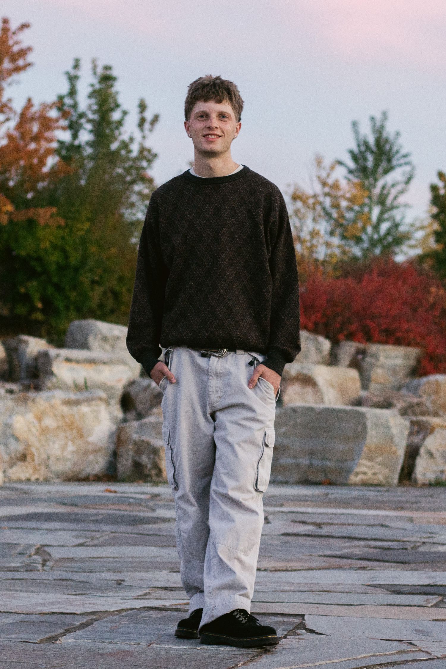 Smiling young man in a dark patterned sweater and gray cargo pants stands on a stone patio with rocks and fall-colored trees.