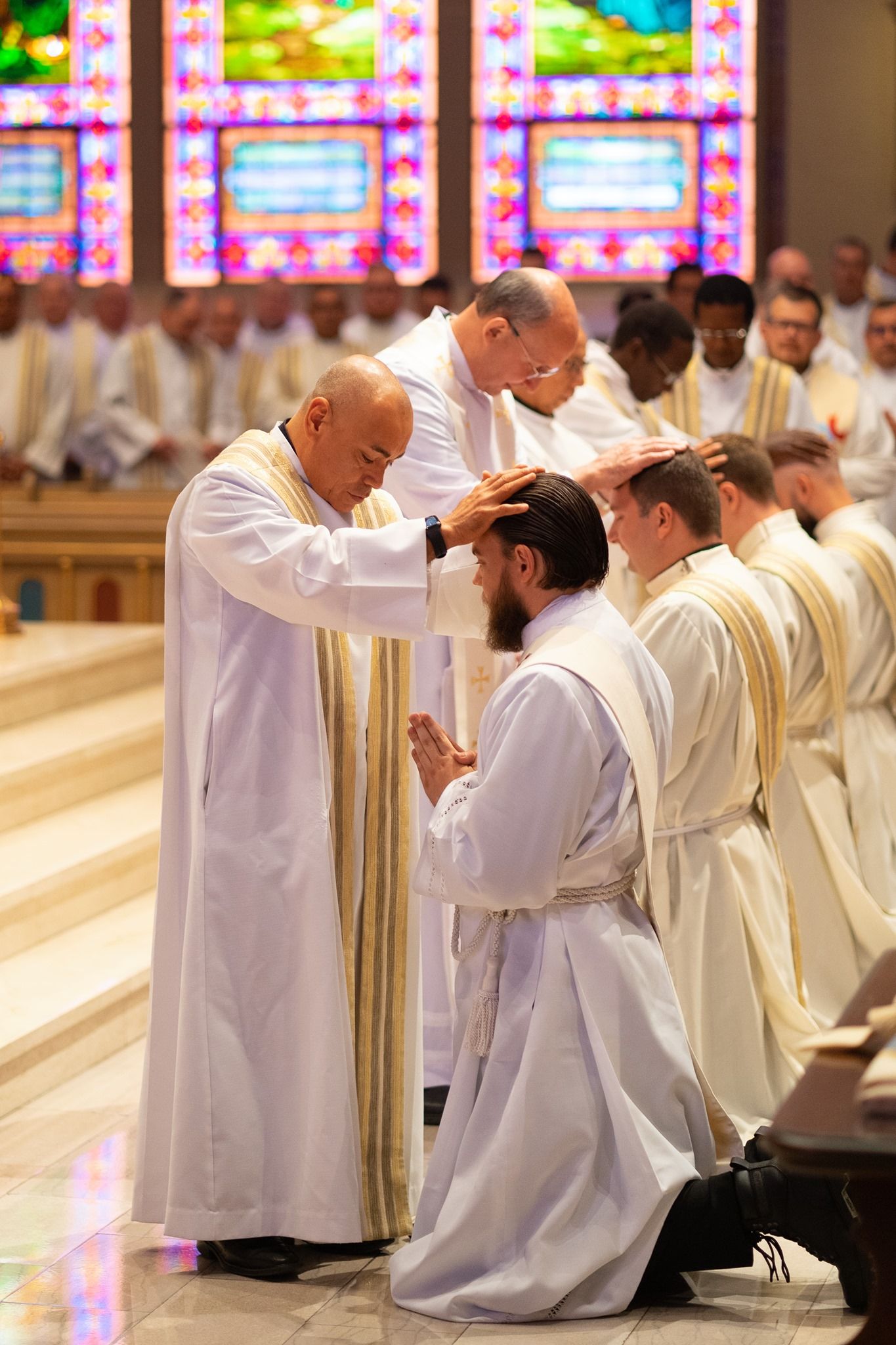 A bishop and priests lay hands on a kneeling man in white vestments during an ordination ceremony in a church with stained glass.