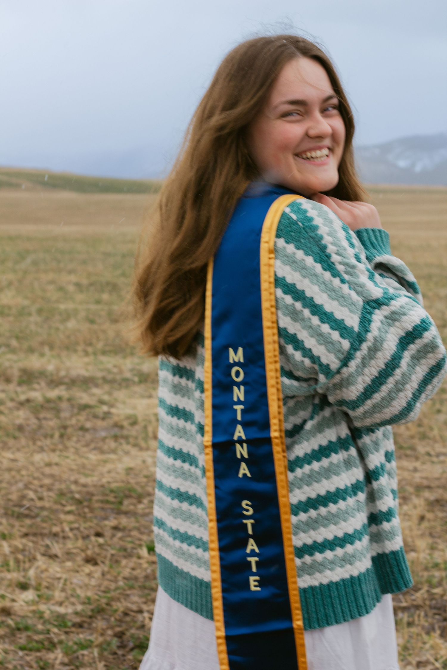 Smiling graduate with long brown hair wears a striped green cardigan and a blue Montana State sash in an open field.