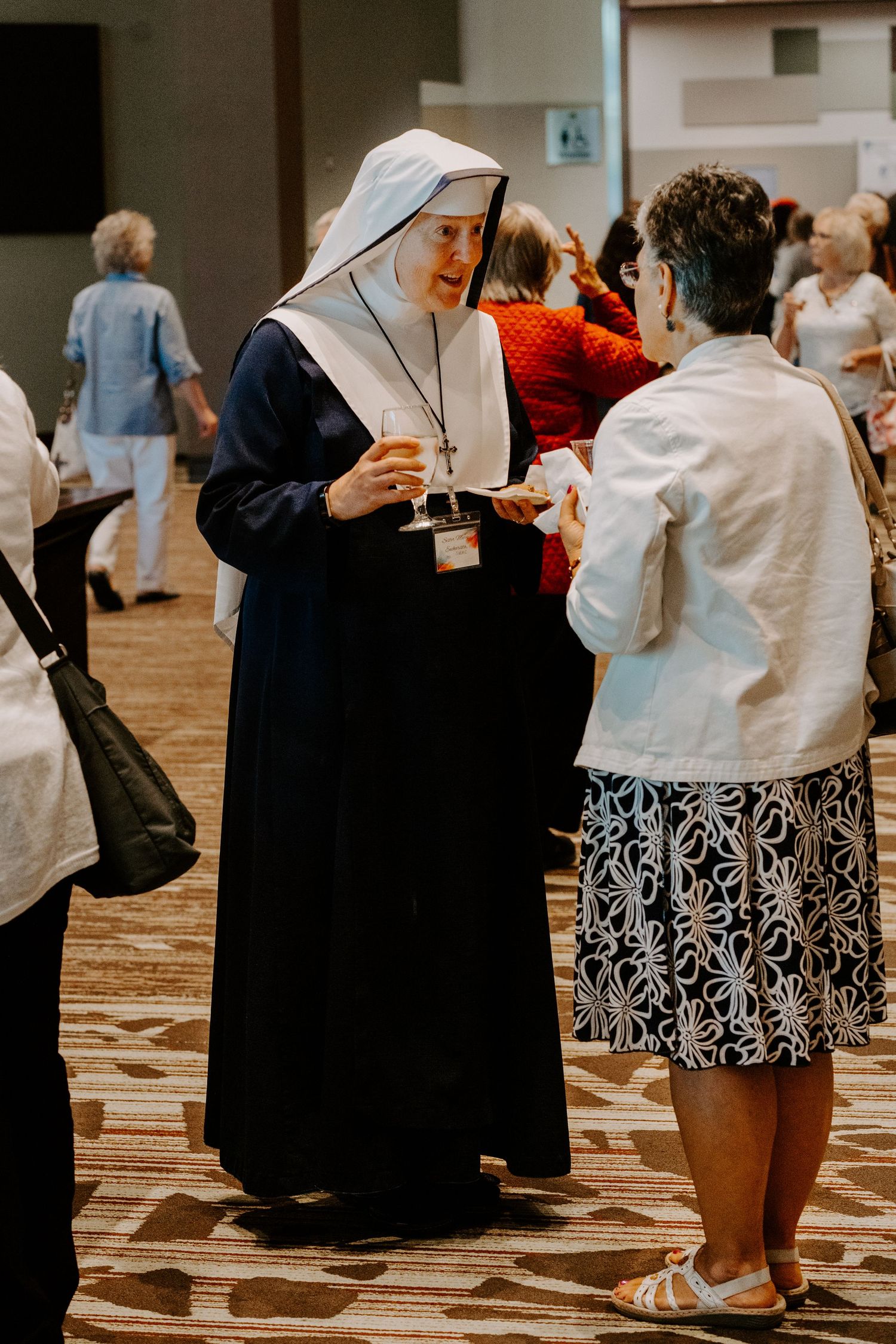 A nun in a white and navy habit holds a drink while chatting with a woman in a floral skirt at a busy indoor event.