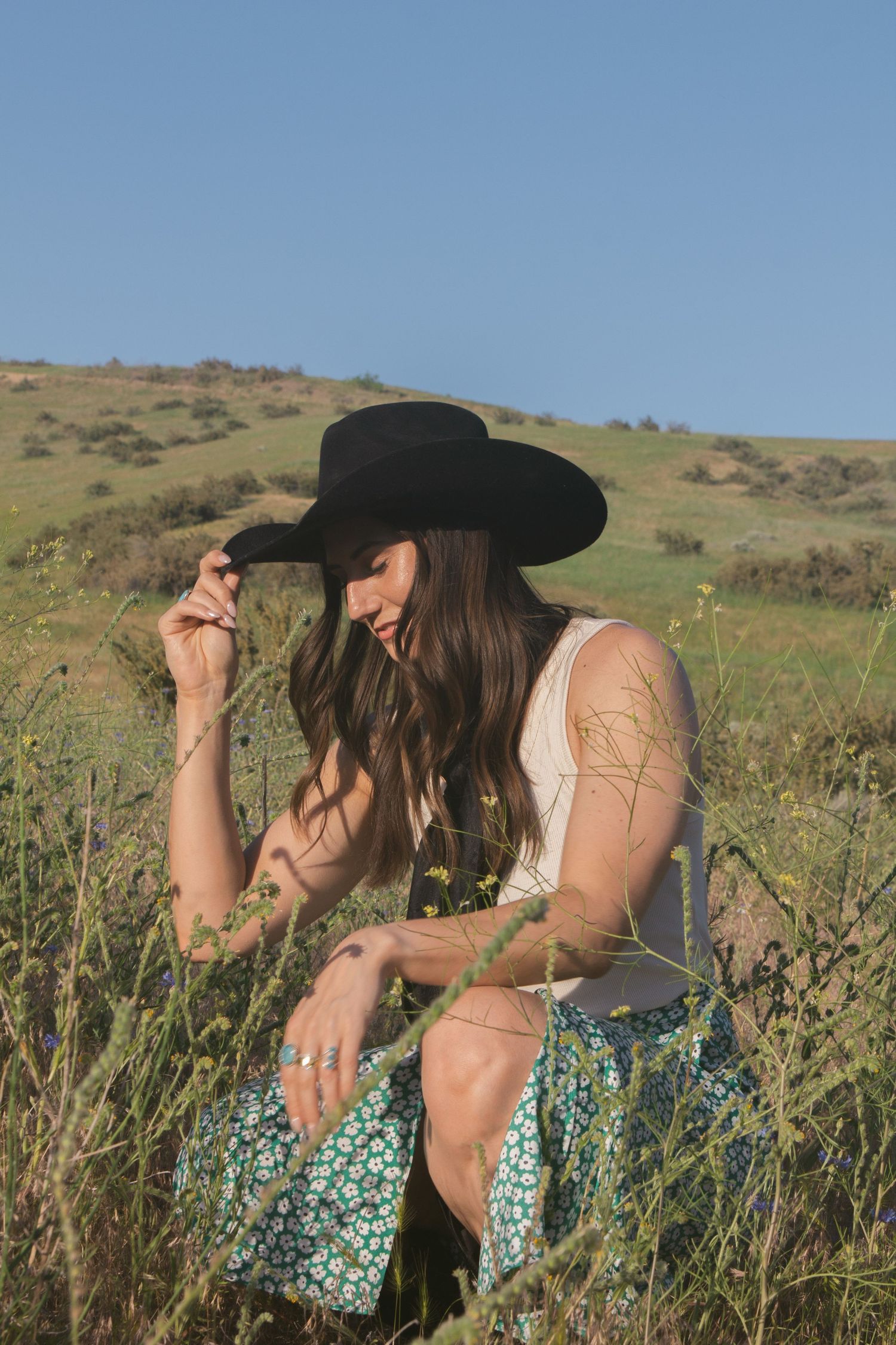 Woman in a black cowboy hat and green floral skirt crouches among tall wild grasses, touching her hat brim on a sunny hillside.