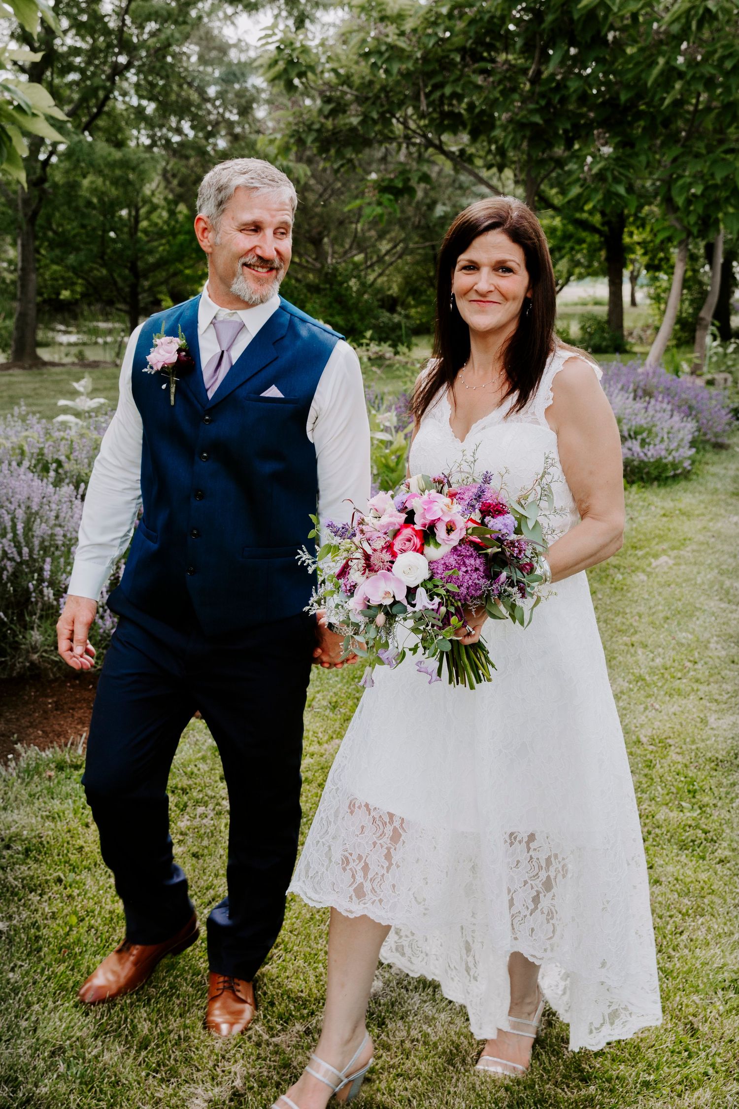 A smiling groom in a navy vest walks hand in hand with a bride in a white lace dress holding a pink bouquet in a garden.