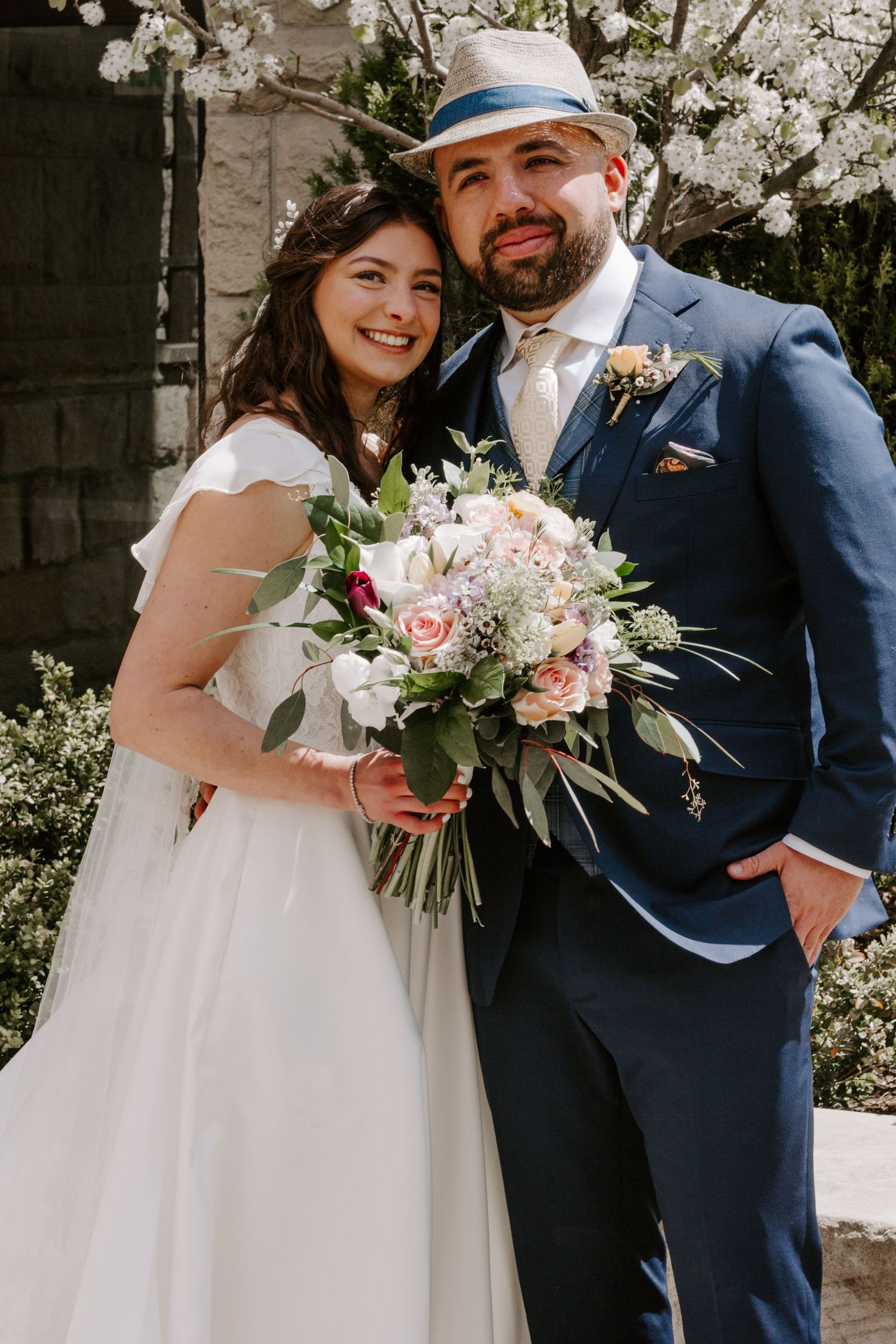 A smiling bride in a flowing white gown and a bearded groom in a navy suit and fedora pose with a bouquet under blossoming trees.