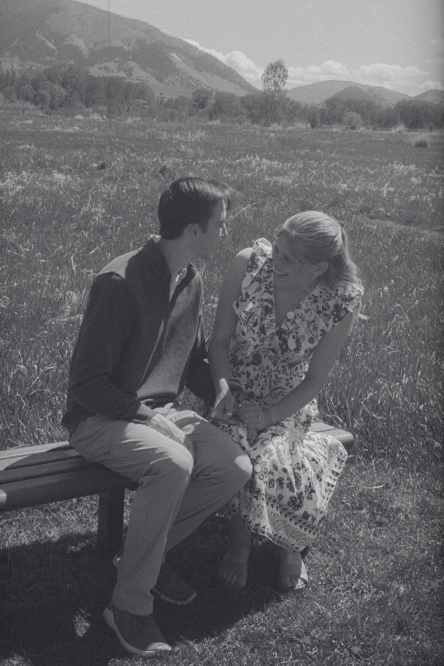 A black and white photo of a man and woman in a floral dress sitting close together on a wooden bench in a grassy mountain field.
