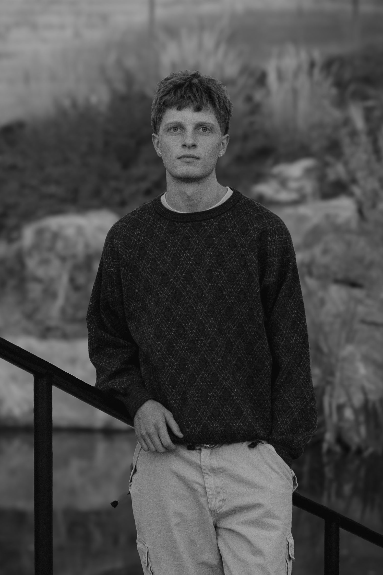 Black and white portrait of a young man in a patterned sweater leaning on a railing beside rocks and a still pond.