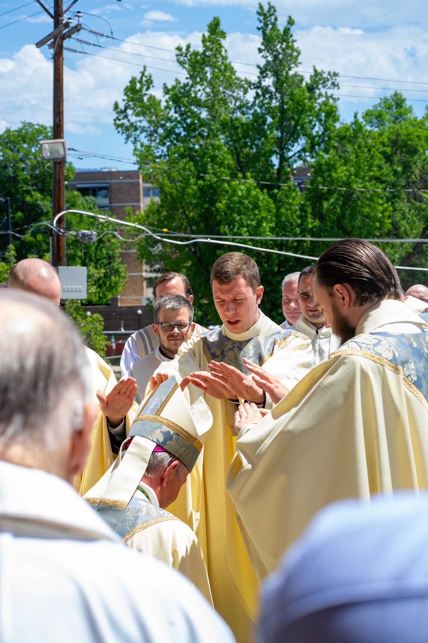 Priests in golden vestments extend their hands over a kneeling man outdoors during an ordination rite beneath green trees and blue sky.
