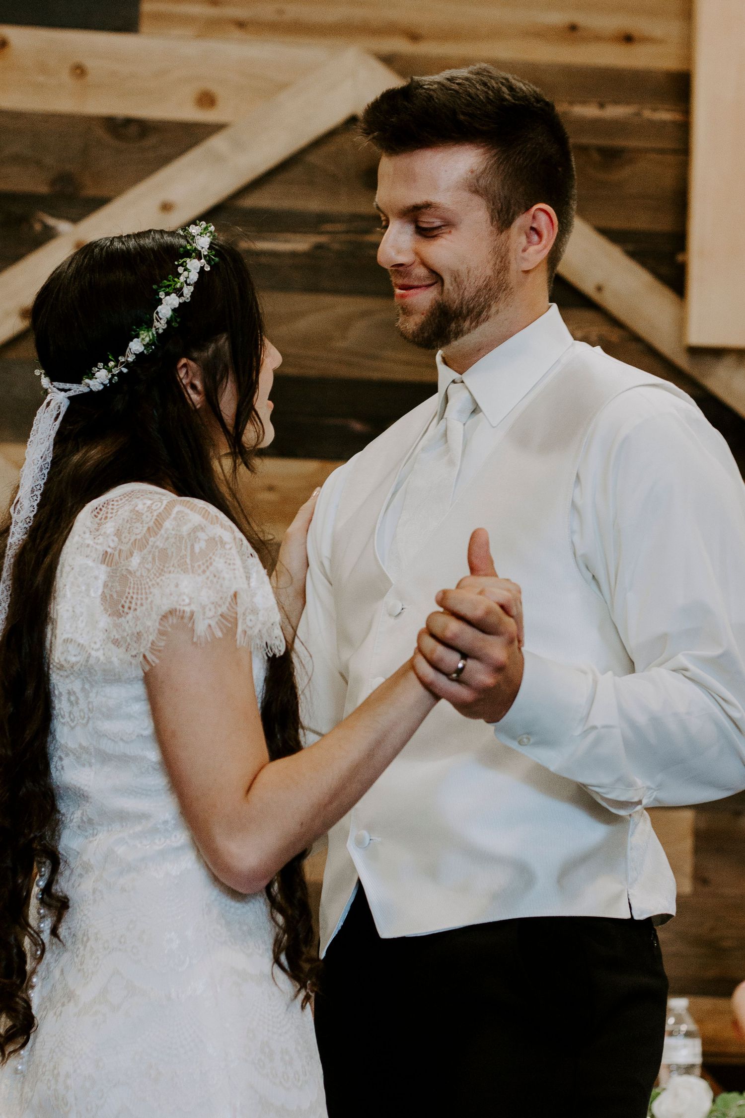 A groom in a white vest smiles at his bride in a lace dress and flower crown as they hold hands during a wedding dance.