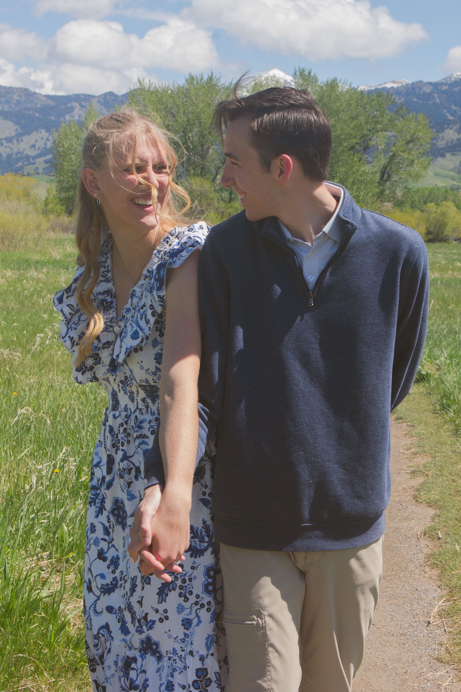Laughing couple holds hands while walking along a dirt path through a green meadow with mountains behind them.