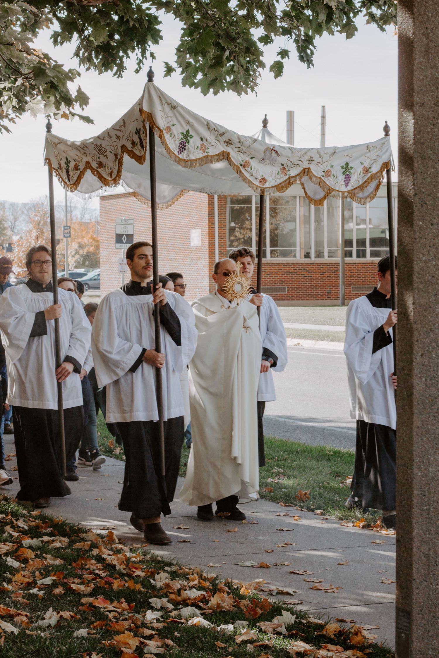 Altar servers and a priest in white vestments walk along a tree-lined sidewalk carrying a canopy in an autumn Eucharistic procession.