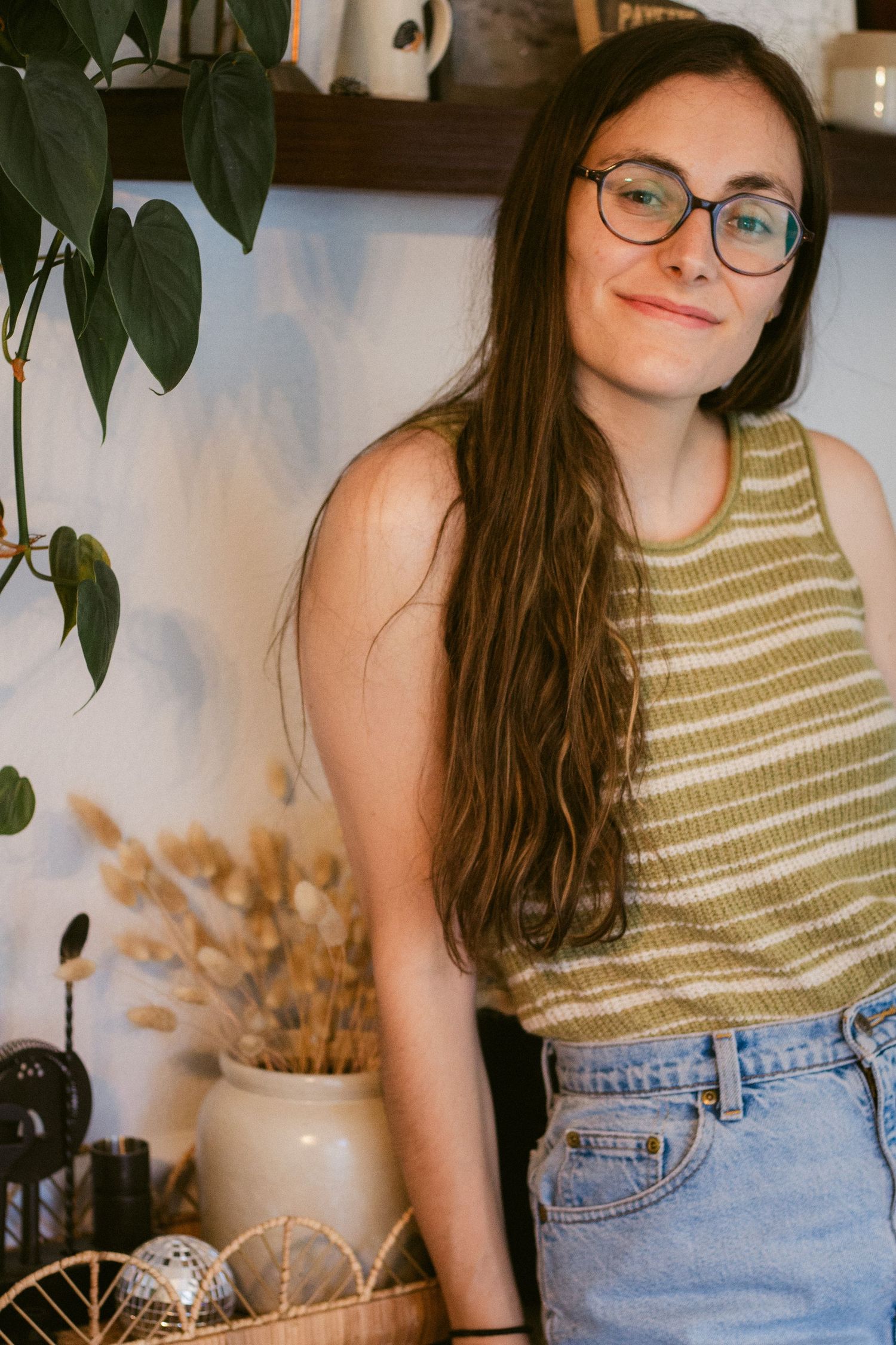 Smiling woman with long brown hair and glasses wears a striped green knit tank top and denim shorts beside plants and dried flowers.