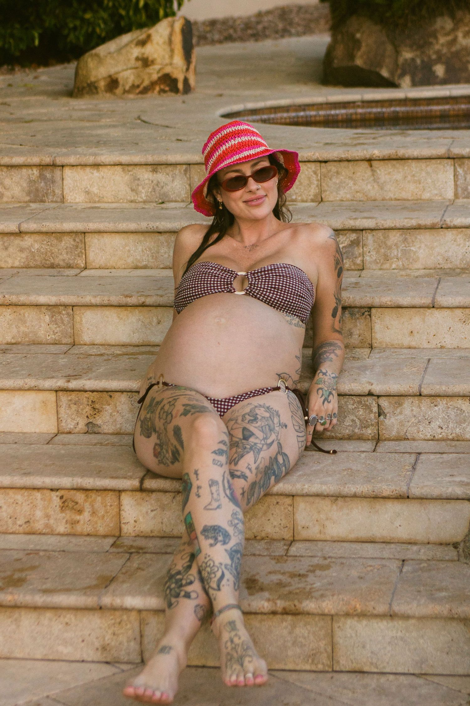 Pregnant woman in a red striped sun hat, sunglasses and brown bikini sits barefoot on sunlit stone steps, her legs covered in tattoos.