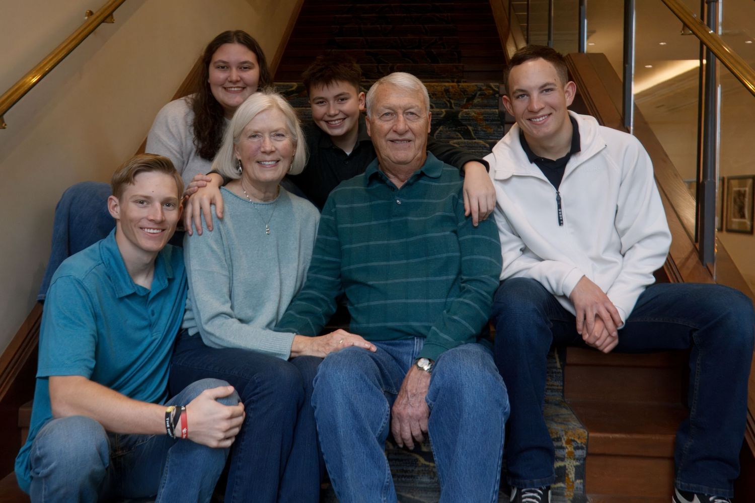 Family portrait of grandparents, young men and women smiling together on a carpeted staircase in a warm indoor setting.