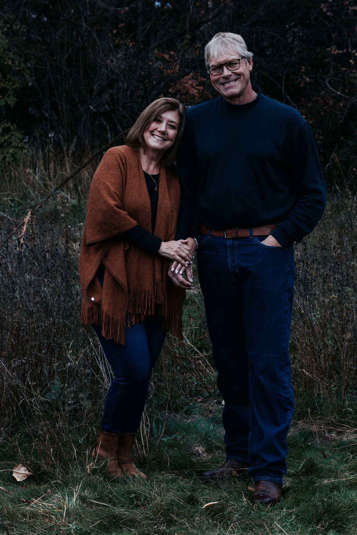 A smiling couple stands arm in arm in a grassy autumn field, she in a rust fringed shawl and he in a dark sweater and jeans.