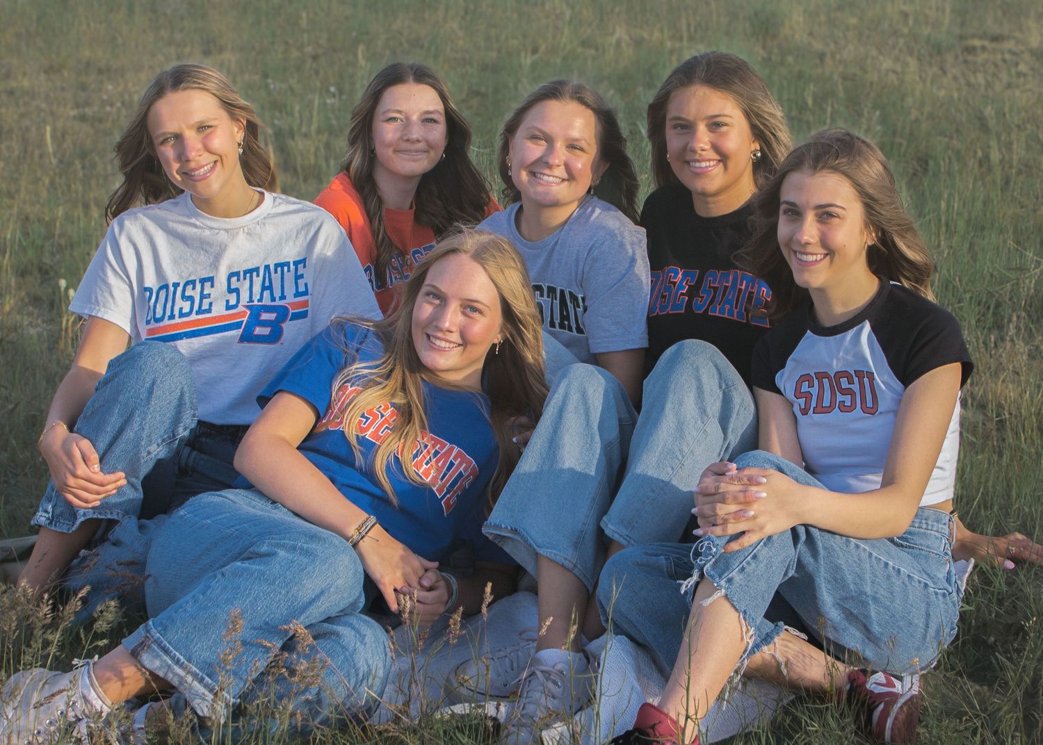 Six smiling young women in Boise State and other college shirts sit close together on the grass during a golden evening light.