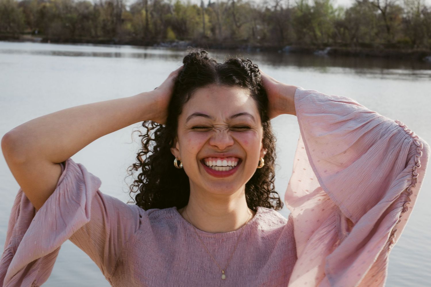 A laughing young woman with curly dark hair holds her hands on her head in a pink blouse beside a calm lake.