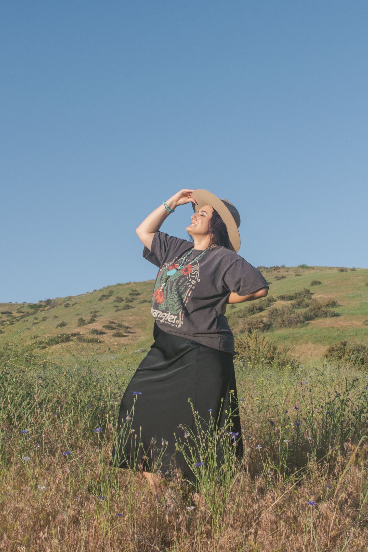 Woman in a graphic tee, black skirt and straw hat gazes upward while standing in a wildflower meadow beneath a clear blue sky.