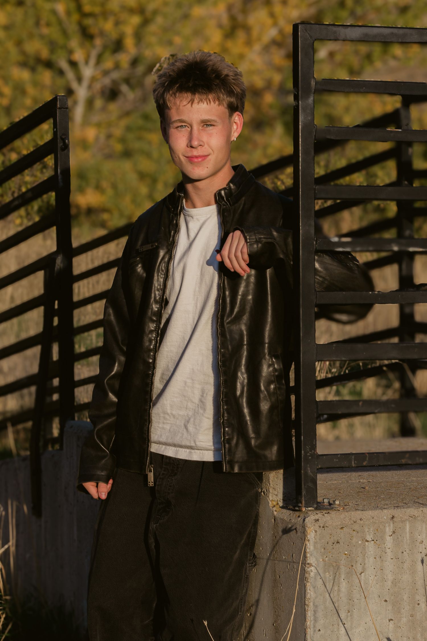 Young man in a black leather jacket and white t-shirt leans against a black metal railing near a concrete wall with autumn trees.