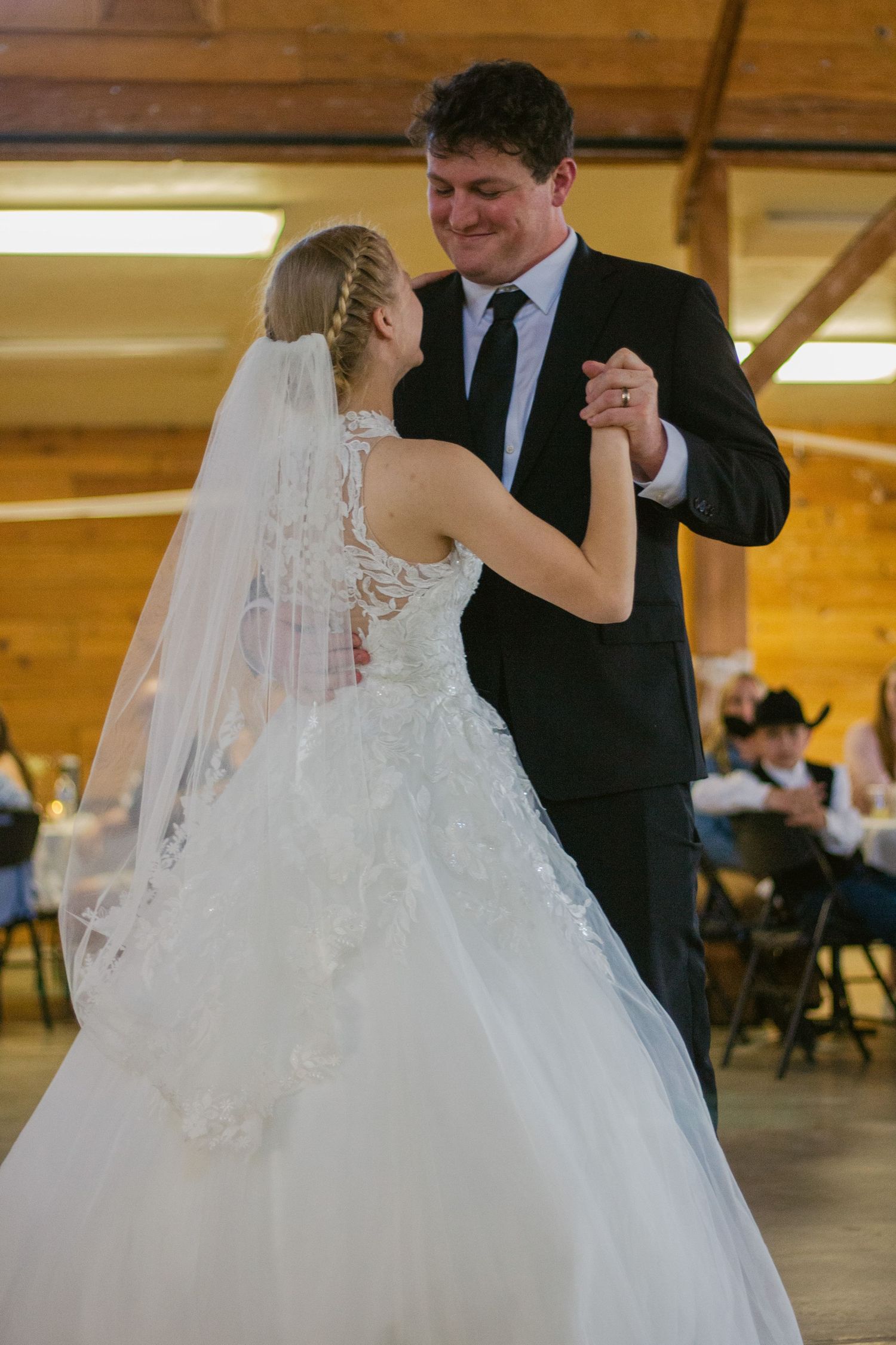 A bride in a lace gown and long veil shares a first dance with a smiling groom in a dark suit in a rustic wooden reception hall.