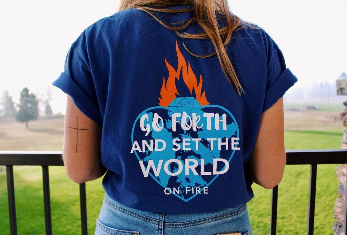 A person wearing a blue t-shirt reading Go Forth and Set the World on Fire with a flaming heart stands facing a green field.