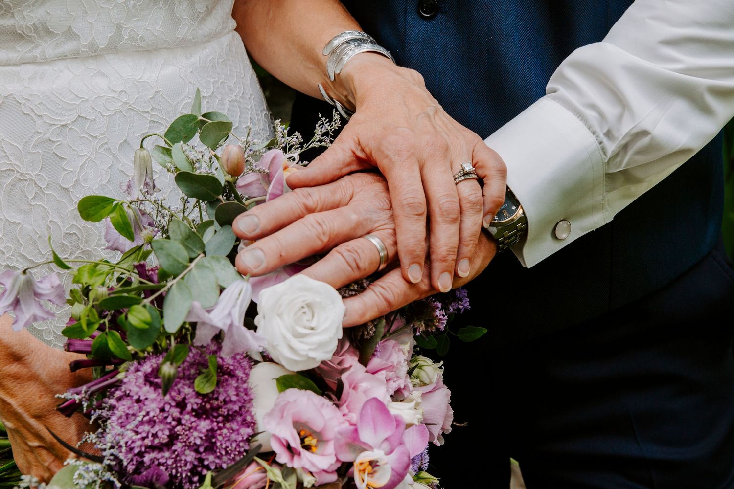 Close-up of a couple's hands with wedding rings resting together above a bouquet of pink, purple and white flowers.