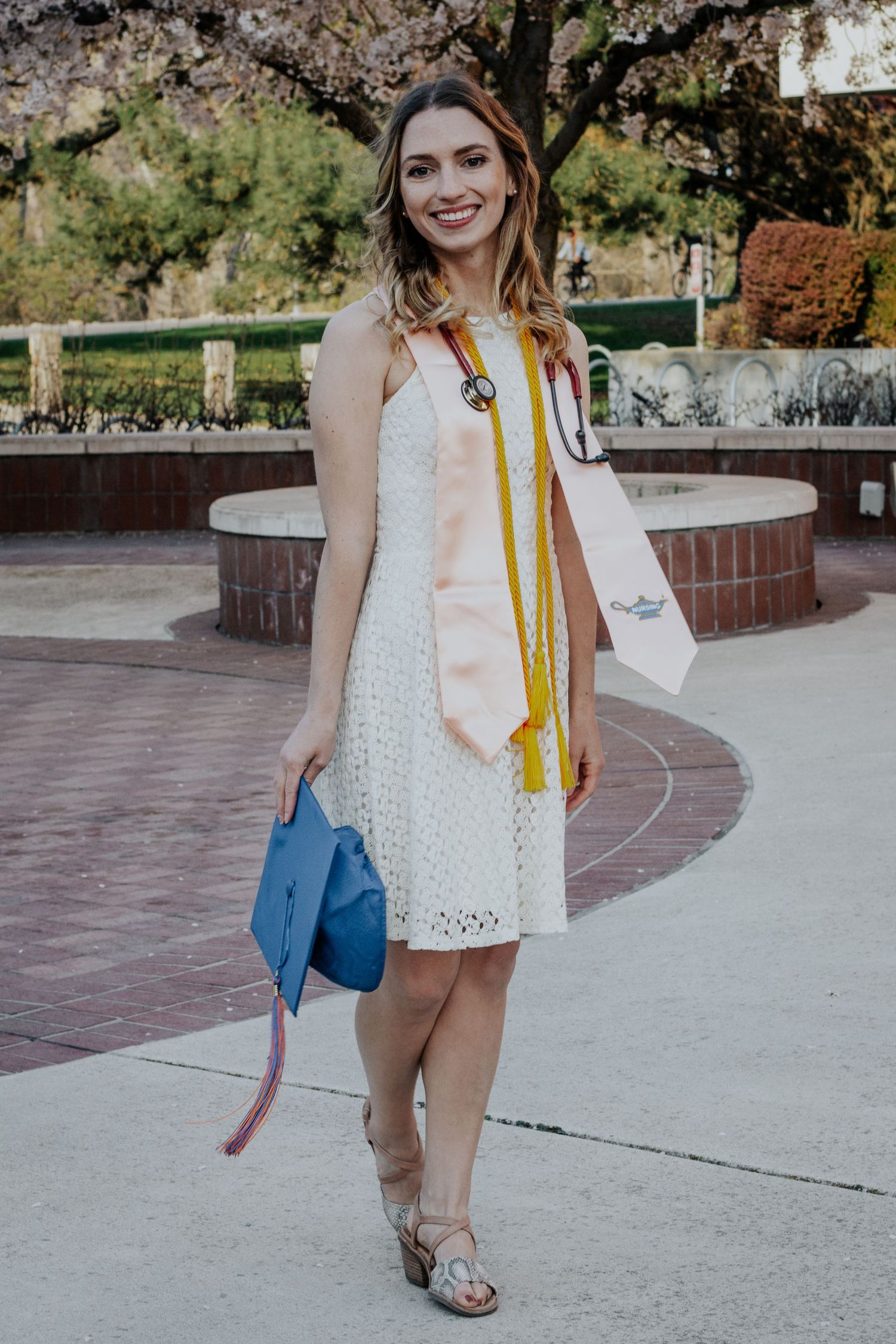 Nursing graduate in a white lace dress with a peach stole, gold cords and stethoscope holds her blue cap beside a brick plaza fountain.